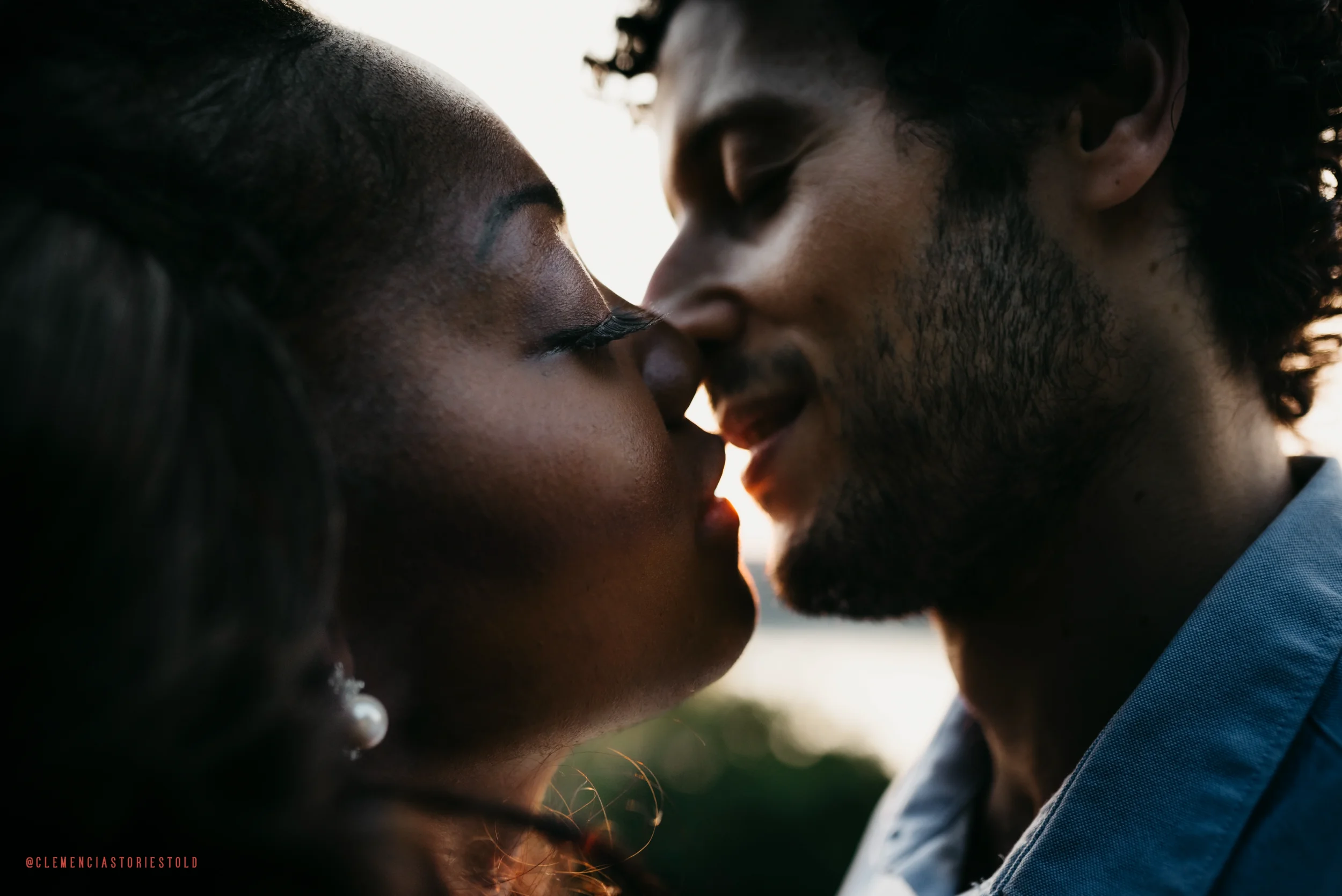 A close-up of a man and woman about to kiss, with faces close together and eyes closed, outdoors during sunset.
