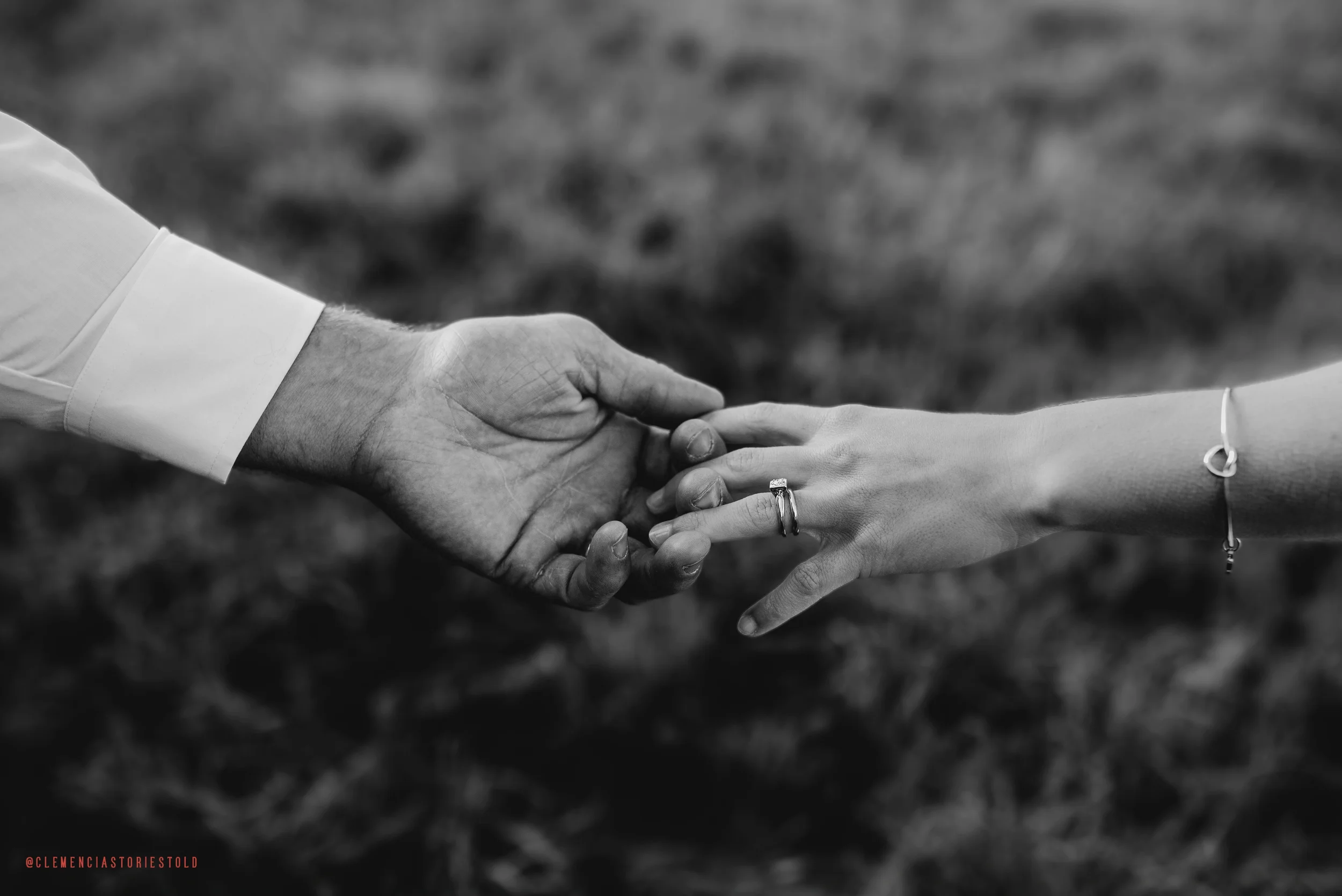 A black-and-white photo of a man and woman holding hands, with the woman's hand showing an engagement ring and a bracelet, and the man's hand appearing larger with visible wrinkles and shirt cuff.