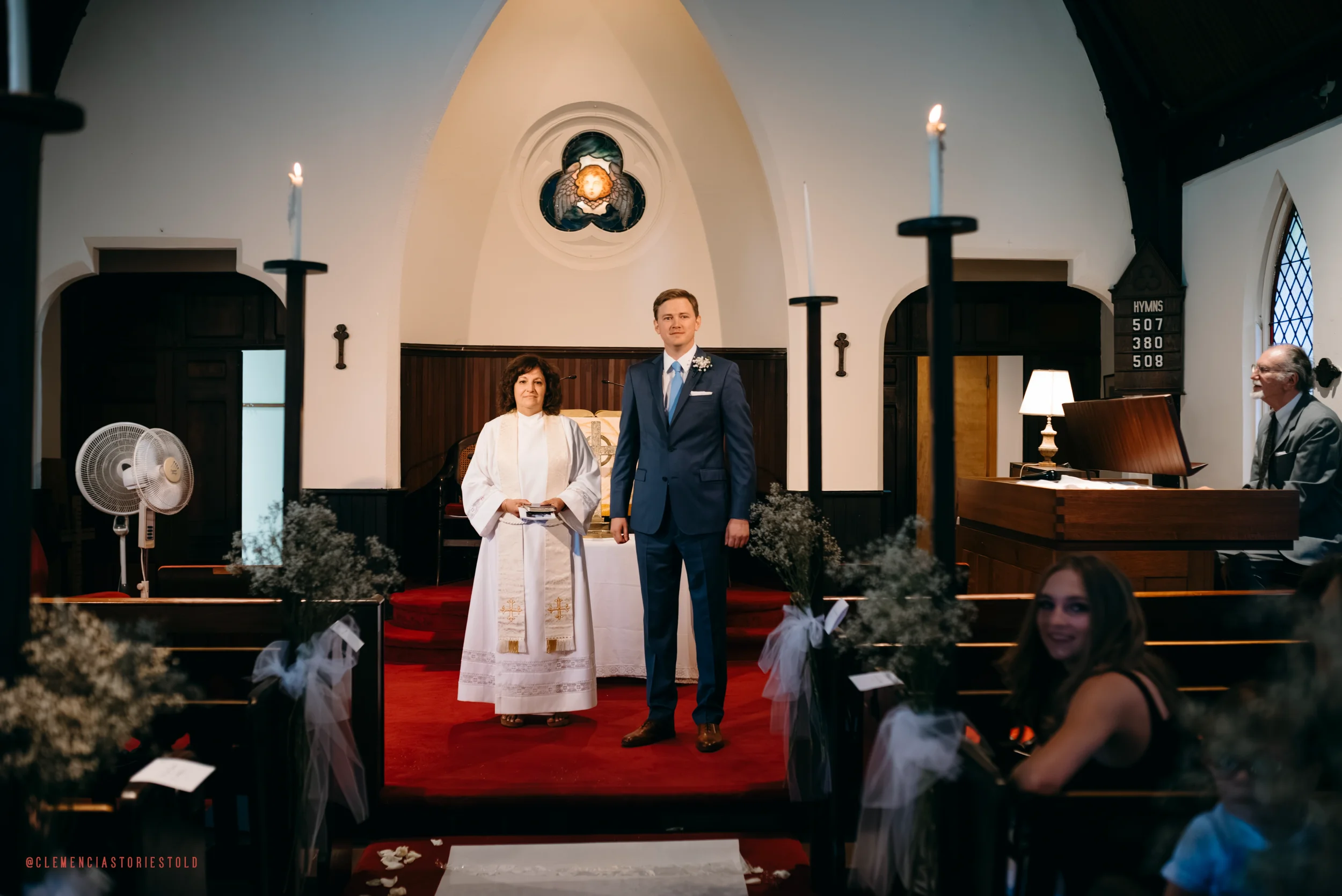 A wedding ceremony inside a church with a bride and groom standing at the altar, officiant woman behind them, pianist to the right, and guests seated in pews decorated with white tulle and flowers.