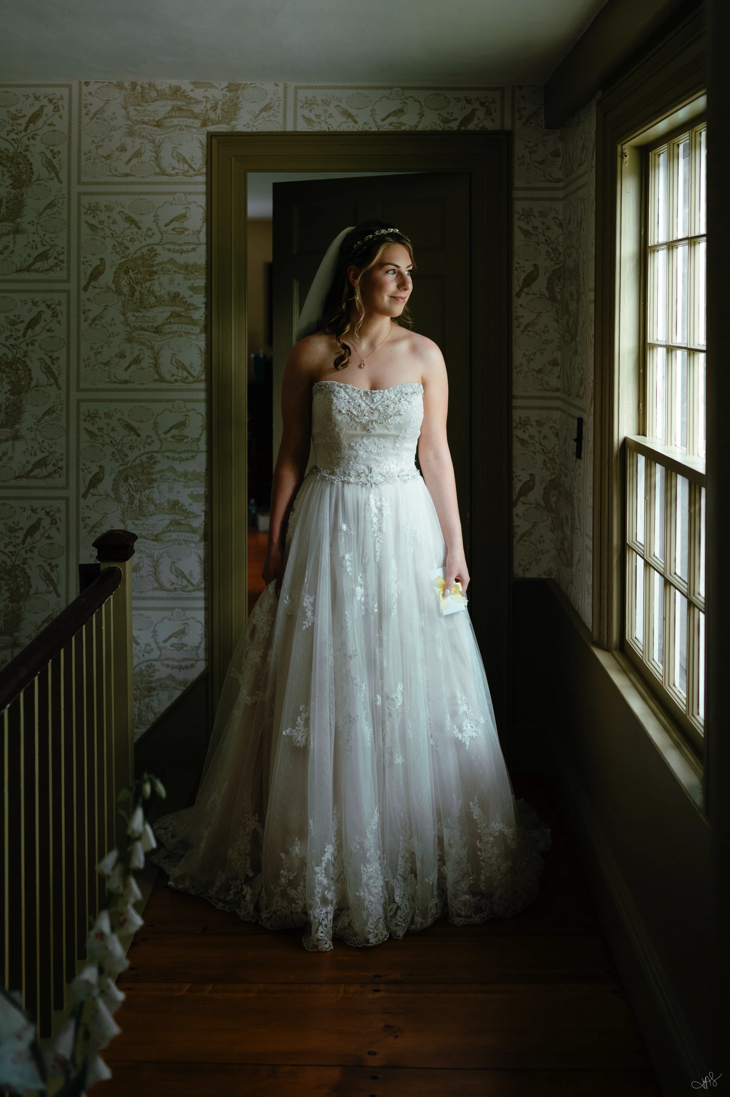 A bride in a strapless white wedding gown with lace details standing in a hallway beside a window, holding a small white bouquet, with soft natural light on her face.