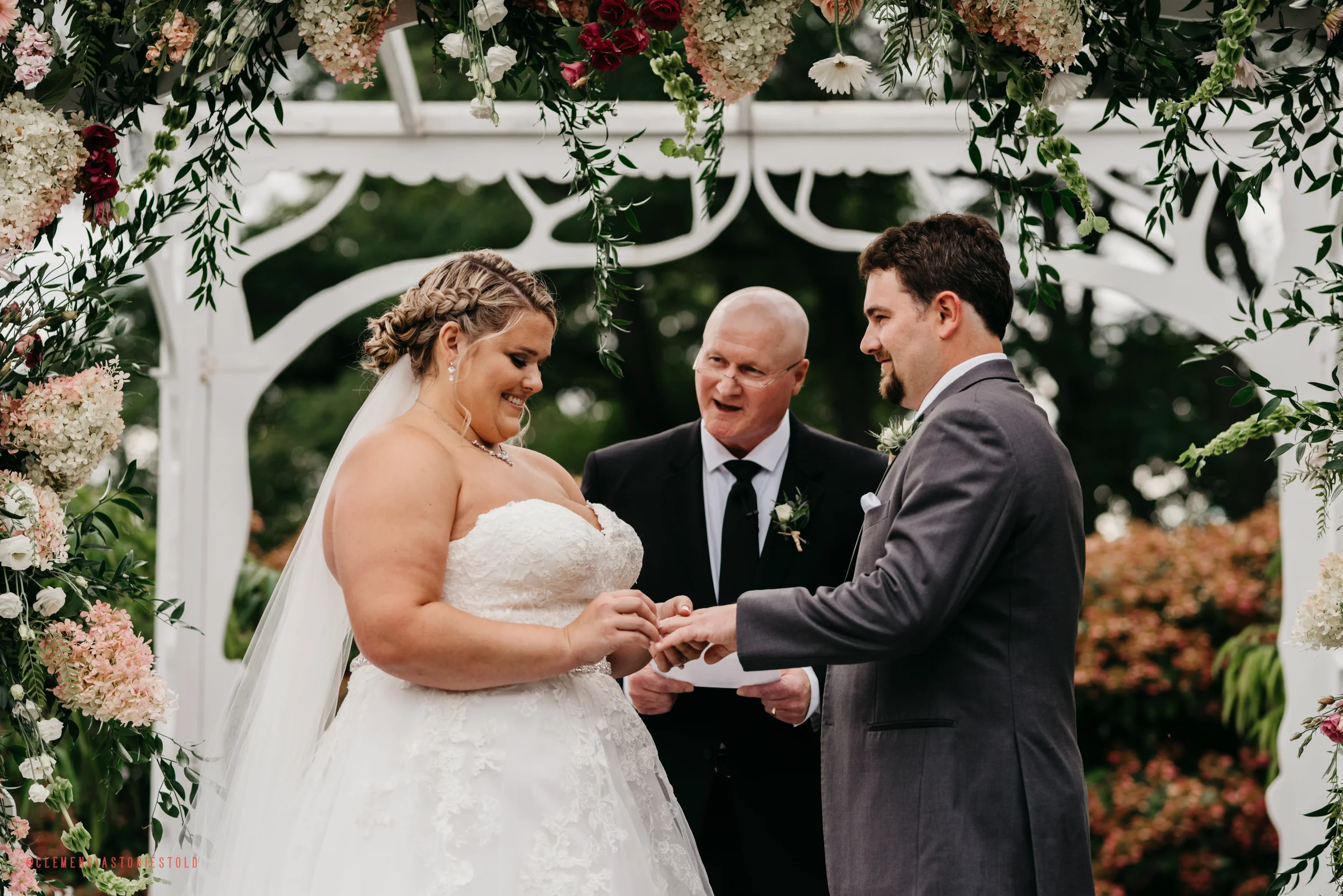 Bride and groom exchanging rings during outdoor wedding ceremony, officiant standing behind them, surrounded by floral arch.
