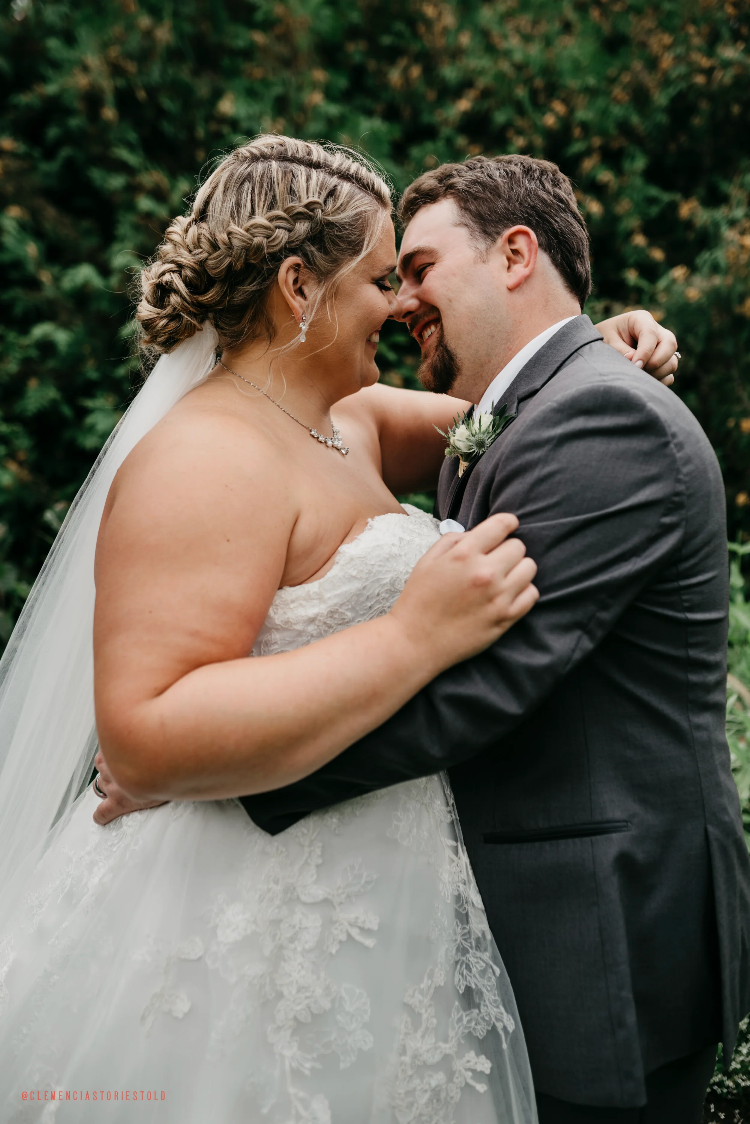 A bride and groom are embracing outdoors, touching foreheads and smiling, with a blurred green background.