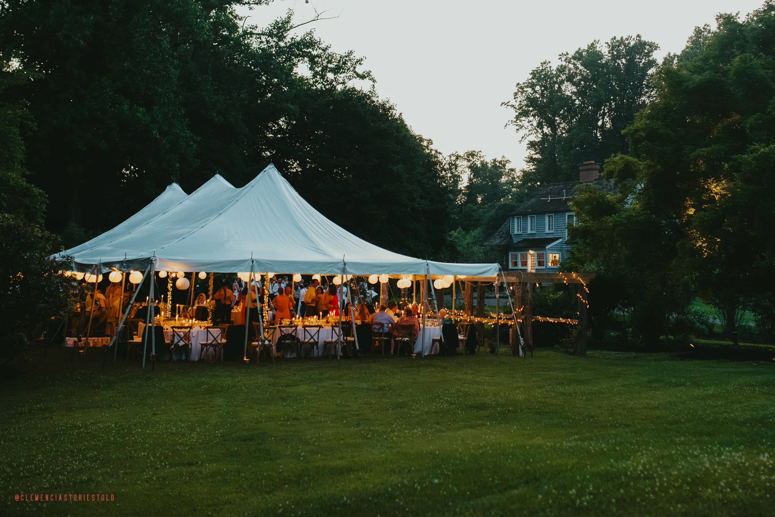 Outdoor evening party under a large white tent with hanging paper lanterns and string lights, surrounded by trees and a house in the background.