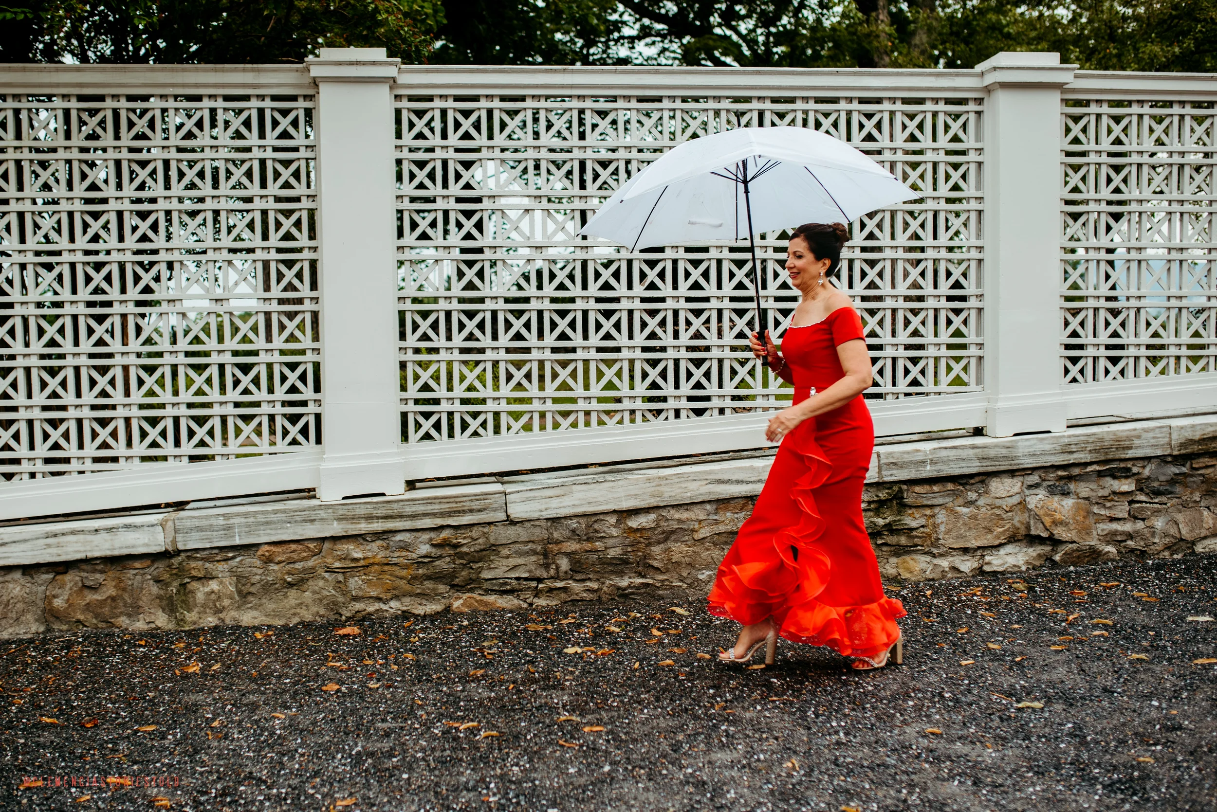 Woman in a red dress holding a white umbrella walking on a gravel path beside a white picket fence.