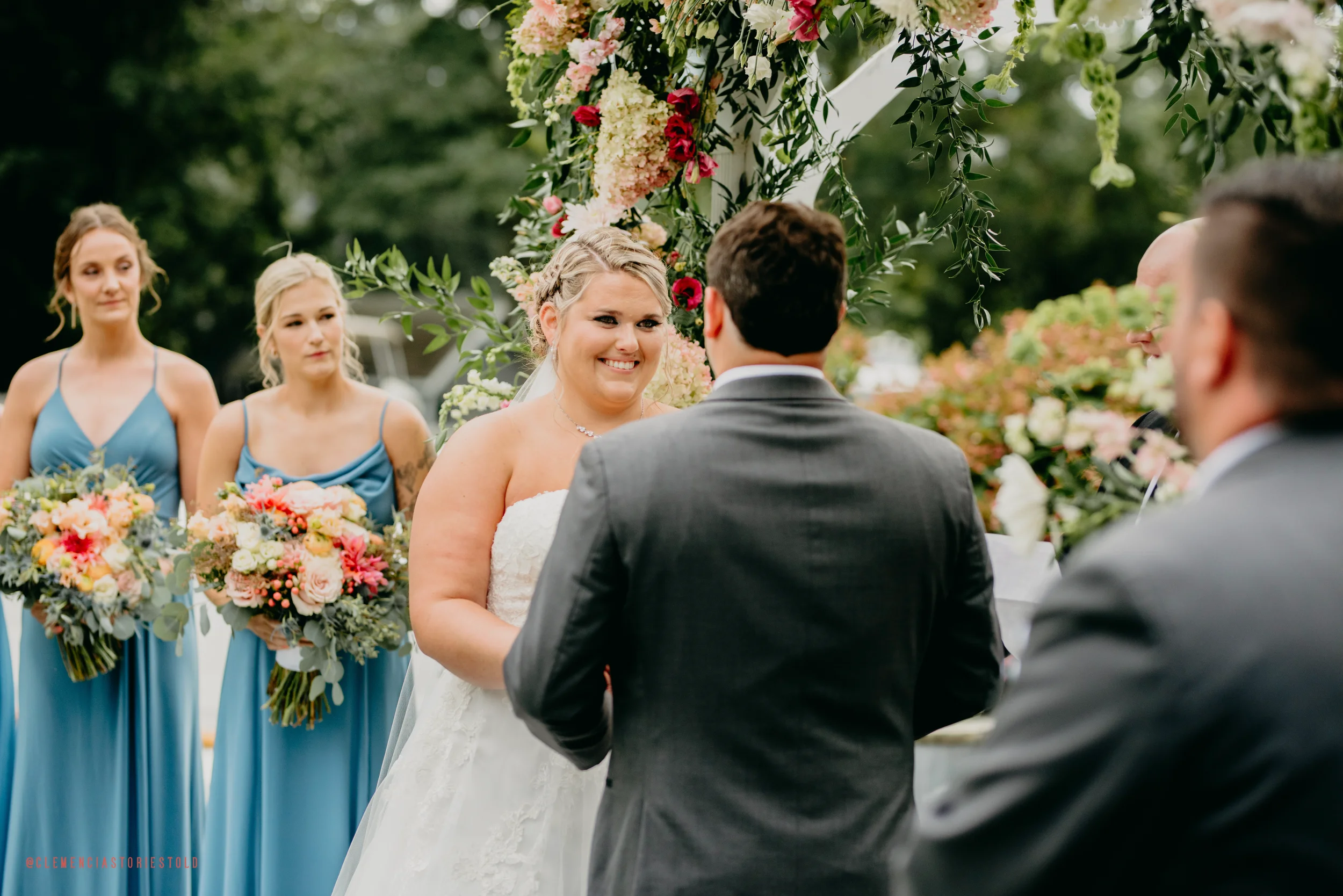 A bride and groom exchanging vows at their outdoor wedding ceremony with bridesmaids holding bouquets standing in the background.