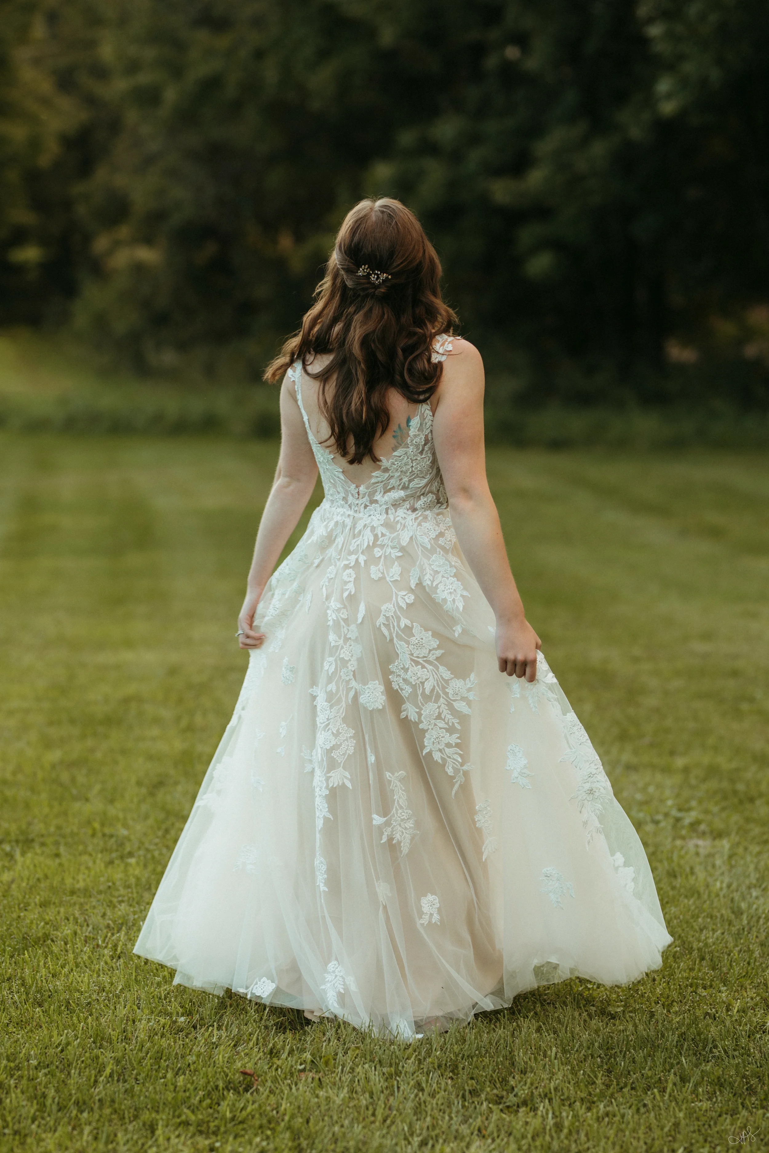 Back view of a woman in a white lace wedding dress standing on grass in a park with trees in the background.