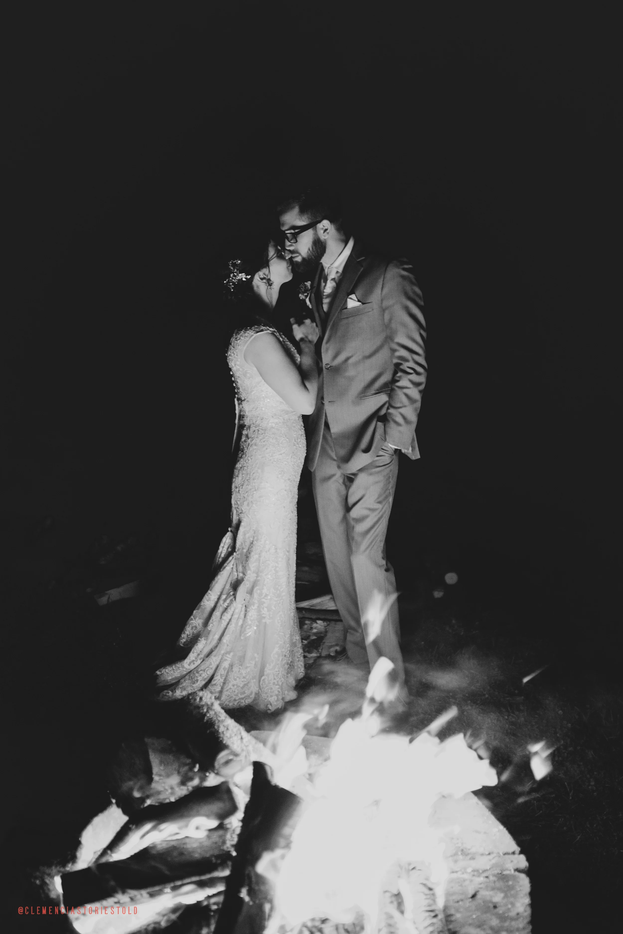 Black and white photo of a bride and groom standing close together by a fire at night, the bride in a lace wedding dress and the groom in a suit.