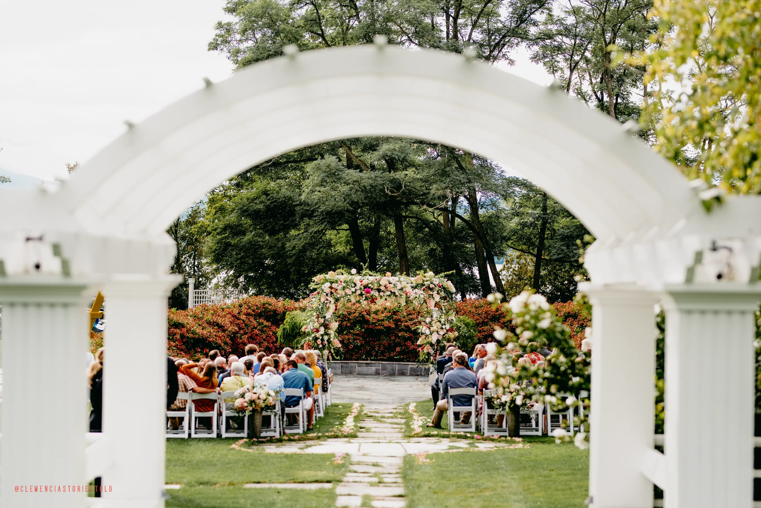 Outdoor wedding ceremony with guests seated on white chairs, floral arrangements, and a large floral arch, viewed through a white archway with trees in the background.