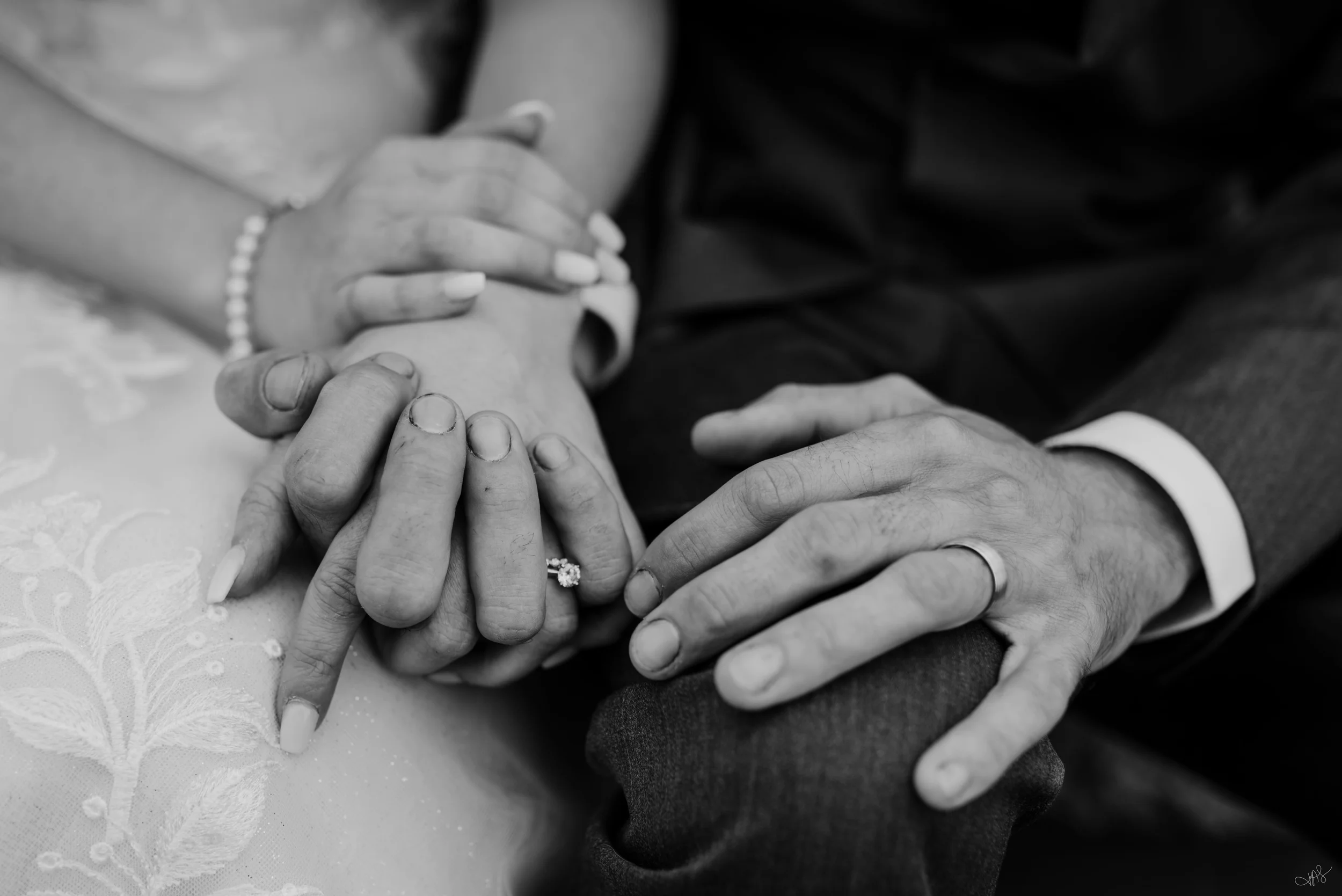Close-up of multiple hands holding a wedding ring, symbolizing a wedding or commitment, with some hands wearing jewelry, on a textured white fabric background.