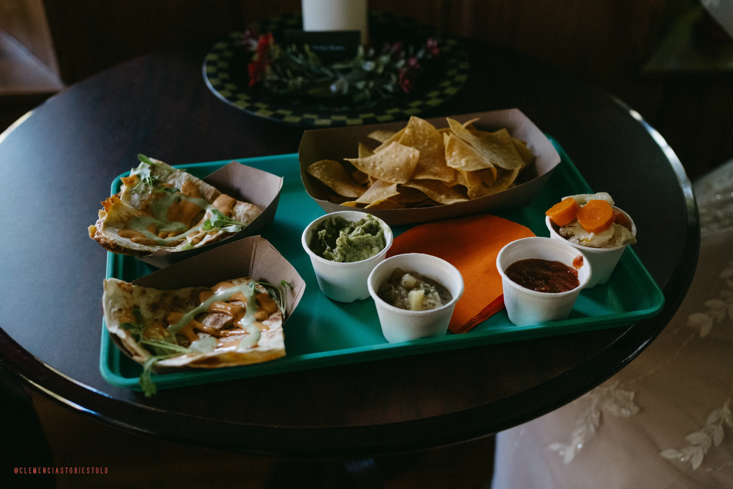 A teal tray holding three slices of pizza with cheese and toppings, a small bowl of guacamole, a small bowl of salsa, a small bowl of refried beans, a small bowl of shredded beef with carrots, a pile of tortilla chips, and a napkin, on a dark round t