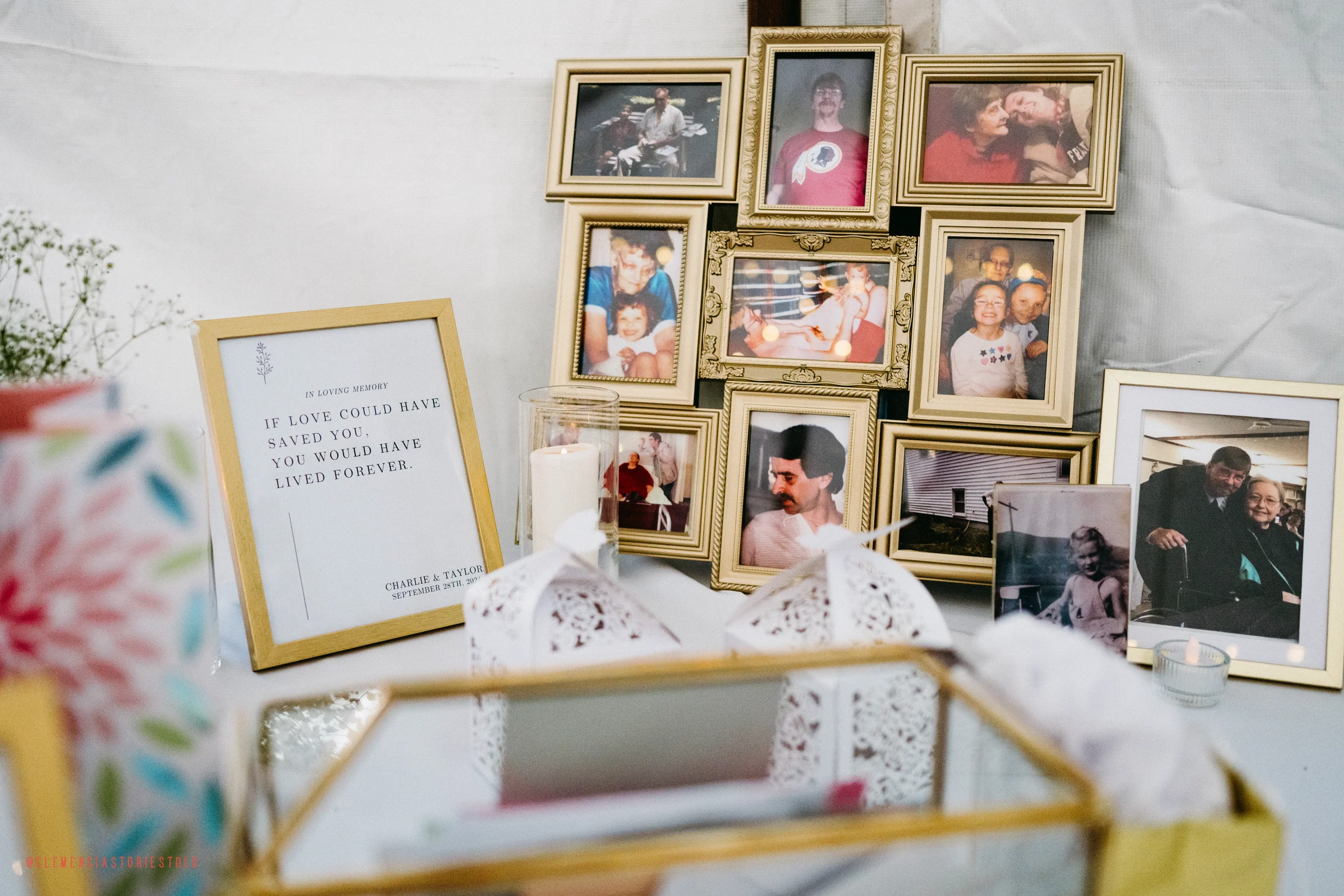 A memorial display with numerous framed photographs of people, a lit candle in a glass holder, a white decorative lace box, and a framed quote that reads, "If love could have saved you, you would have lived forever," with the names Charlie & Taylor a