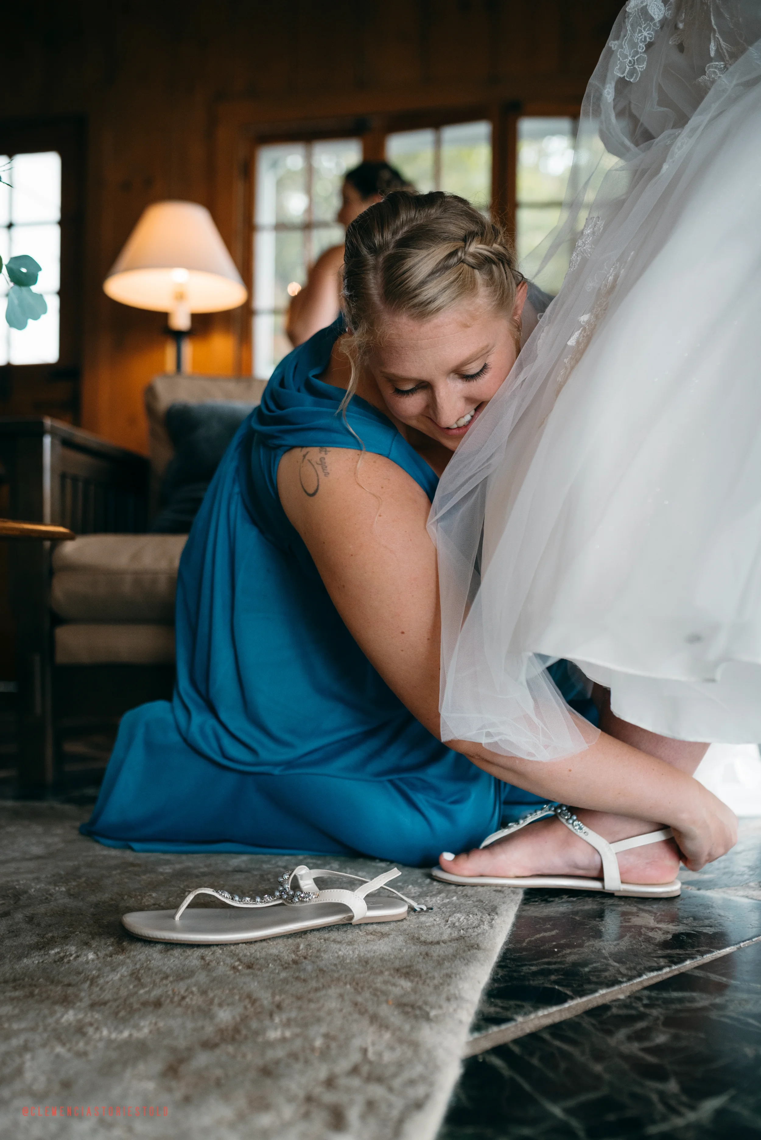 Woman in a blue dress helping a bride with her wedding shoes in a cozy, wood-paneled room with large windows and a lamp in the background.