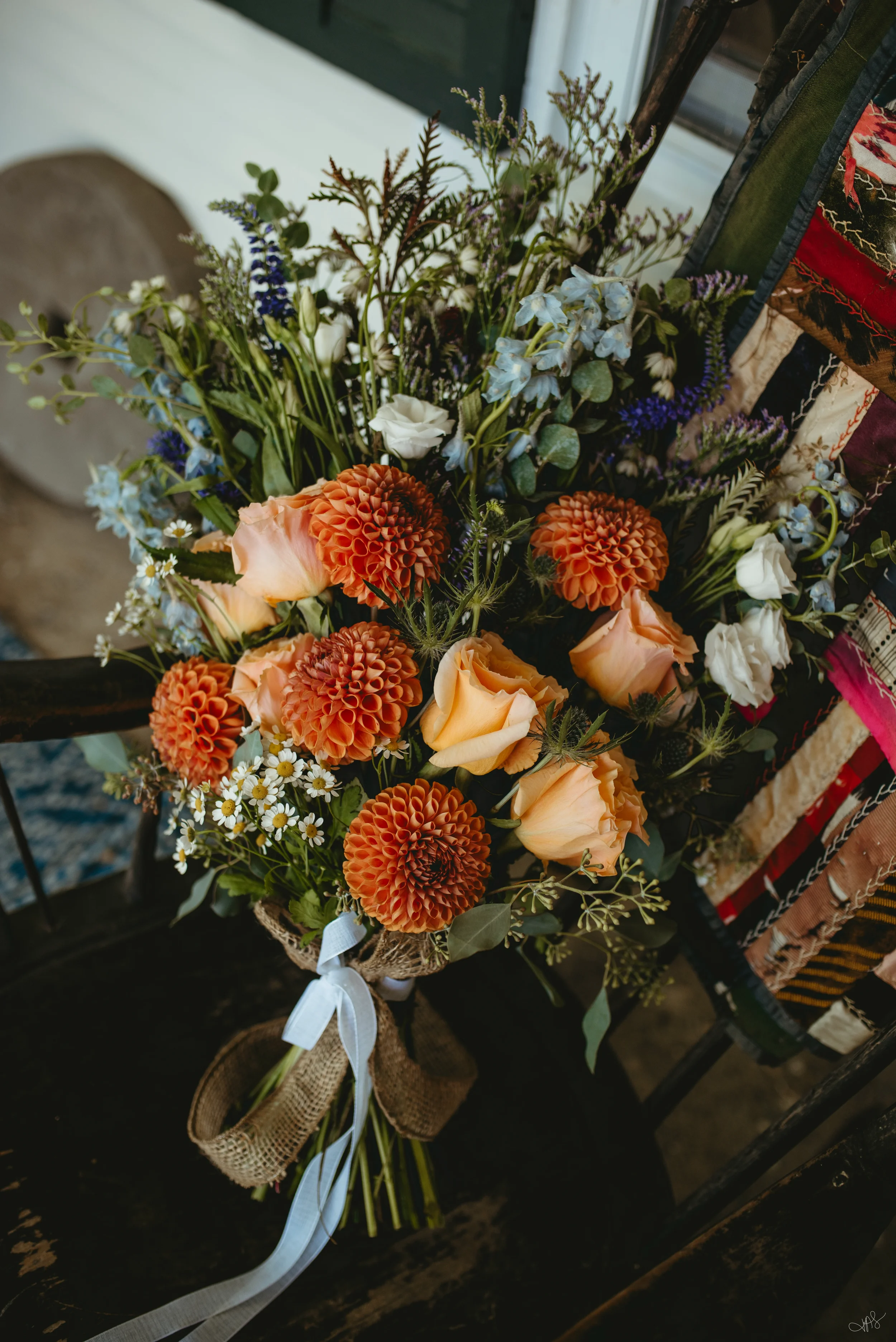 A bouquet of flowers including orange dahlias, cream roses, and various greenery, tied with a white ribbon, resting on a rustic surface.