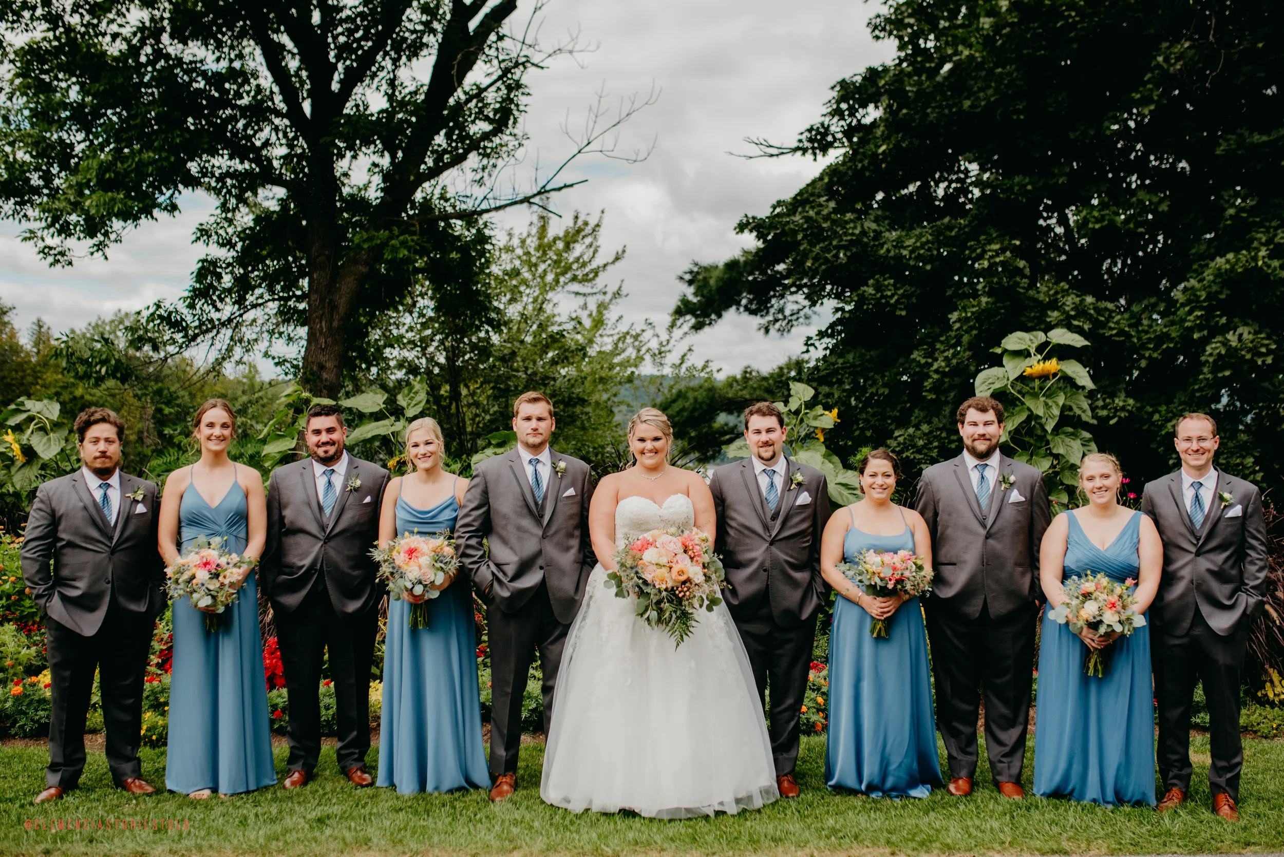 A wedding party standing outdoors on grass with trees and flowers in the background, featuring a bride in a white gown and nine attendants in suits and blue dresses.
