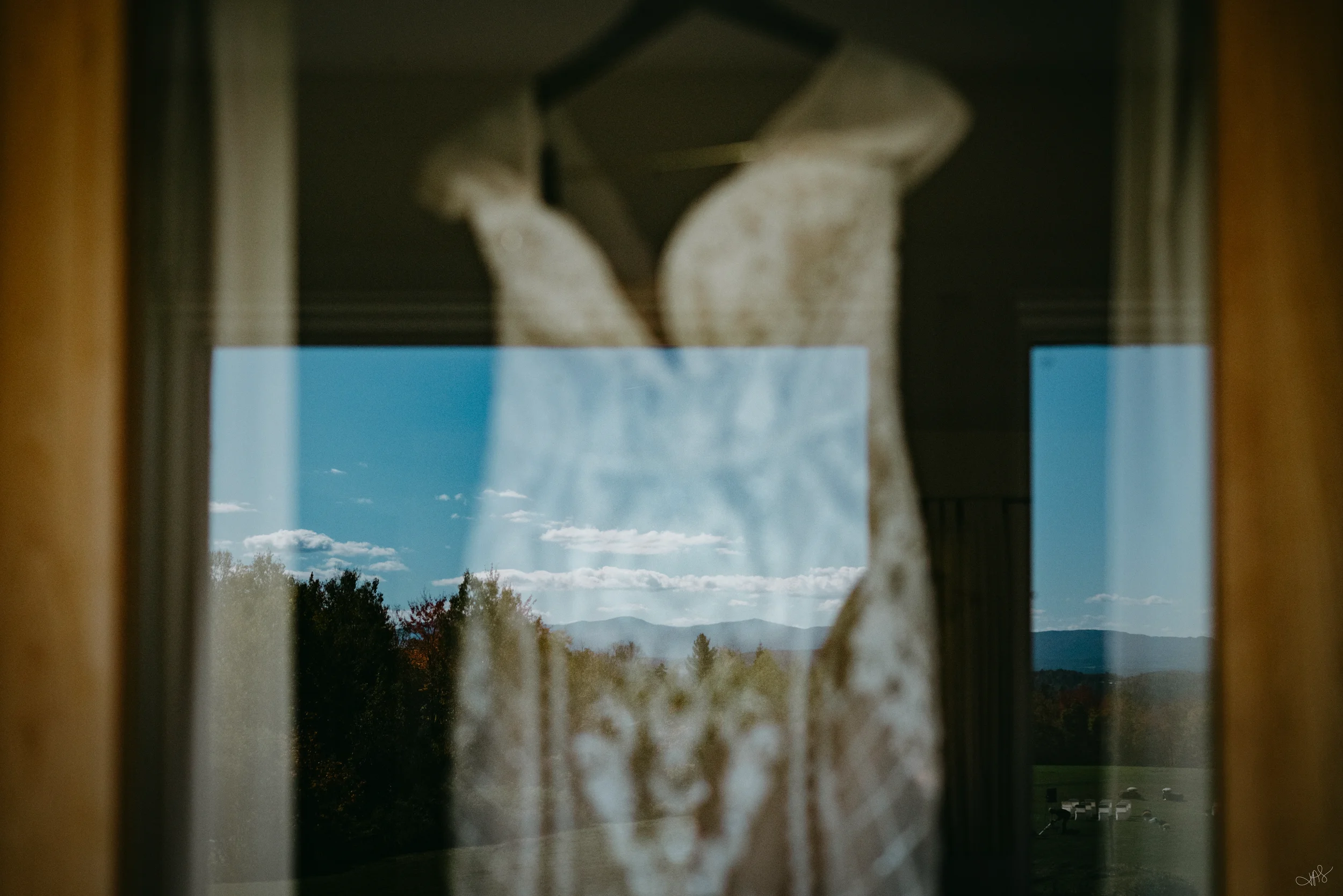 A window with lace curtains viewed from inside, with a landscape of trees, hills, and a cloudy blue sky visible outside.