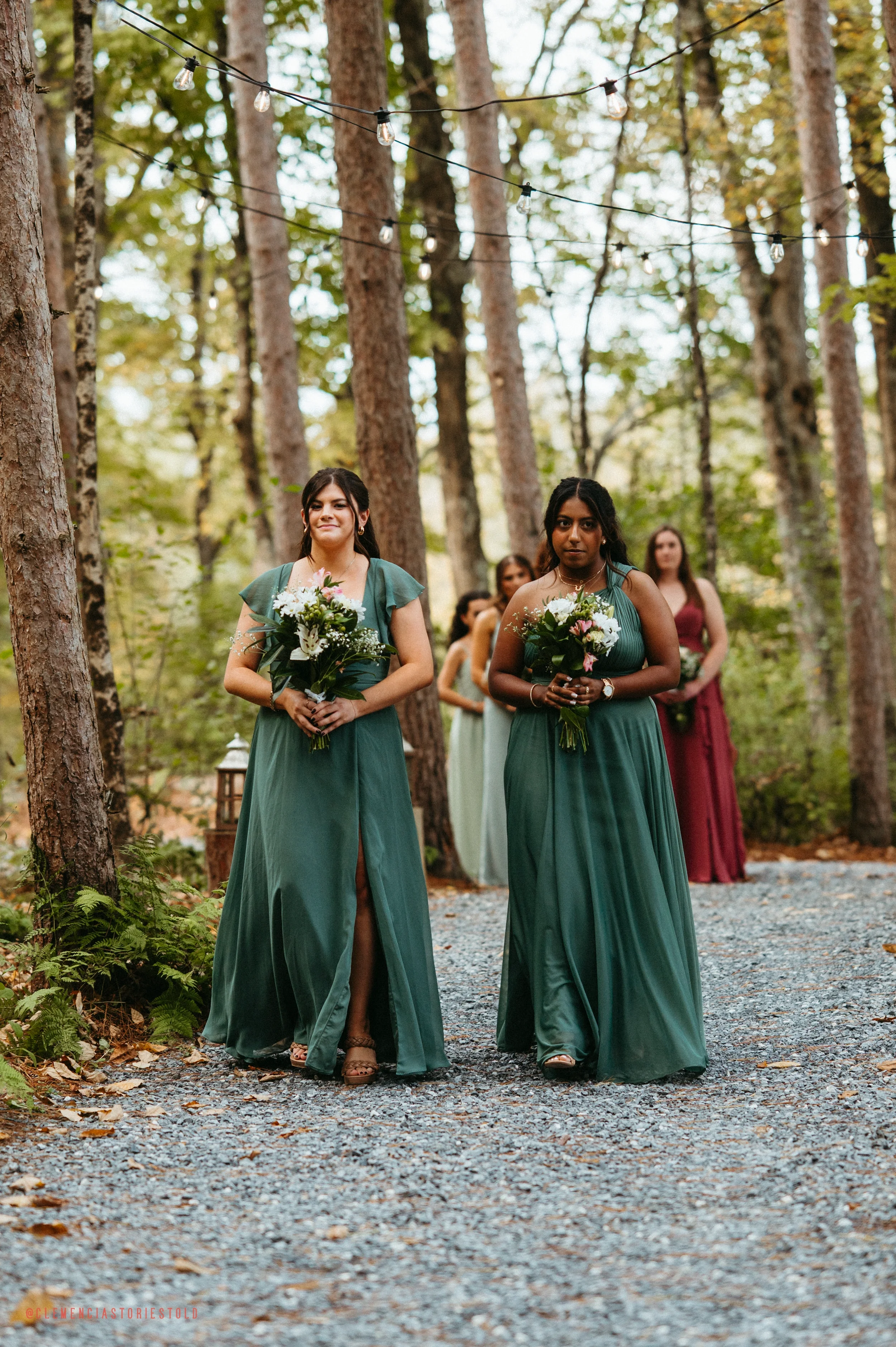 A group of women dressed in teal and burgundy gowns holding bouquets, walking on a gravel path through a wooded area during an outdoor wedding ceremony.