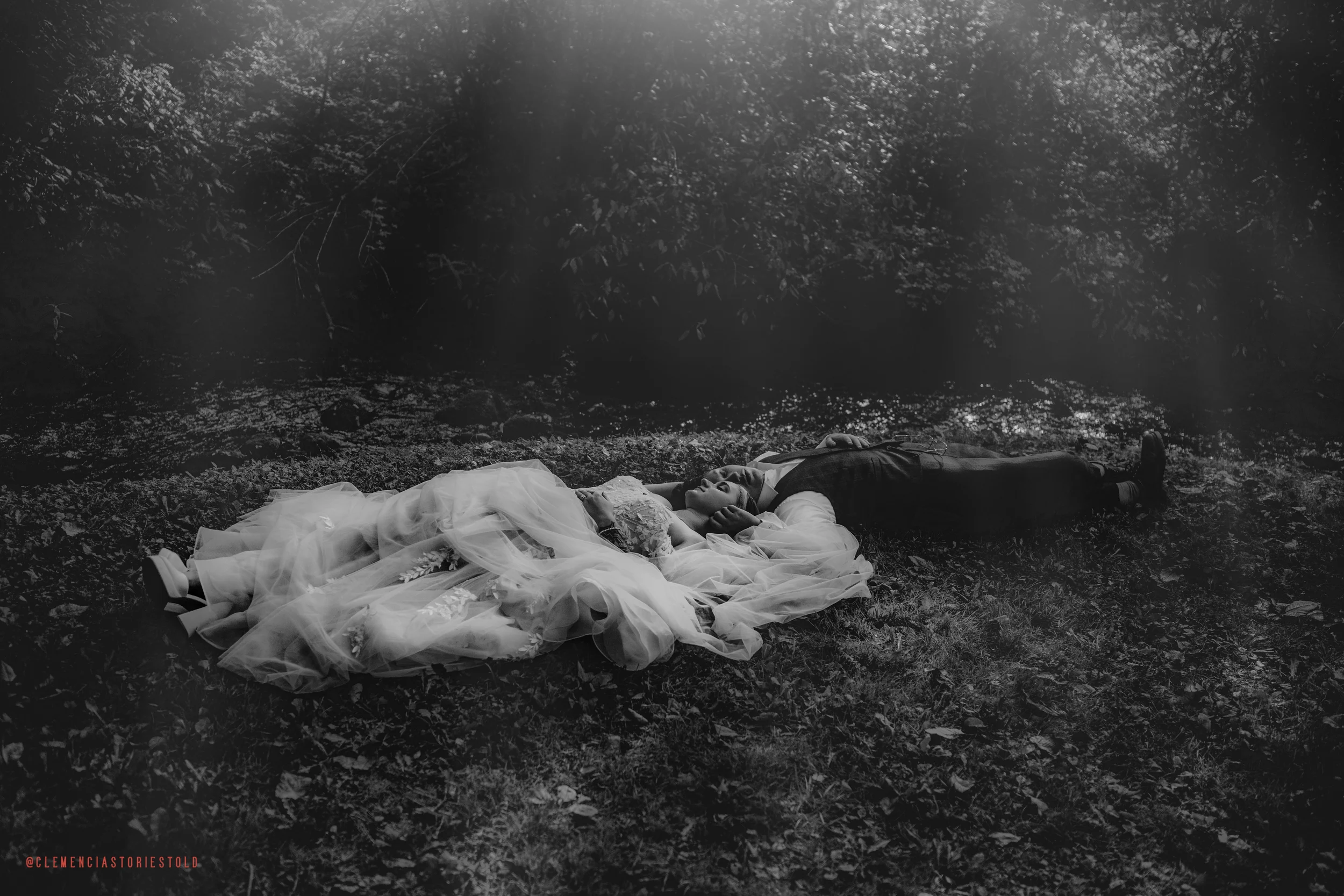 A bride in a wedding dress and veil lying on a grassy ground while a groom in a suit lies beside her in a wooded outdoor setting.