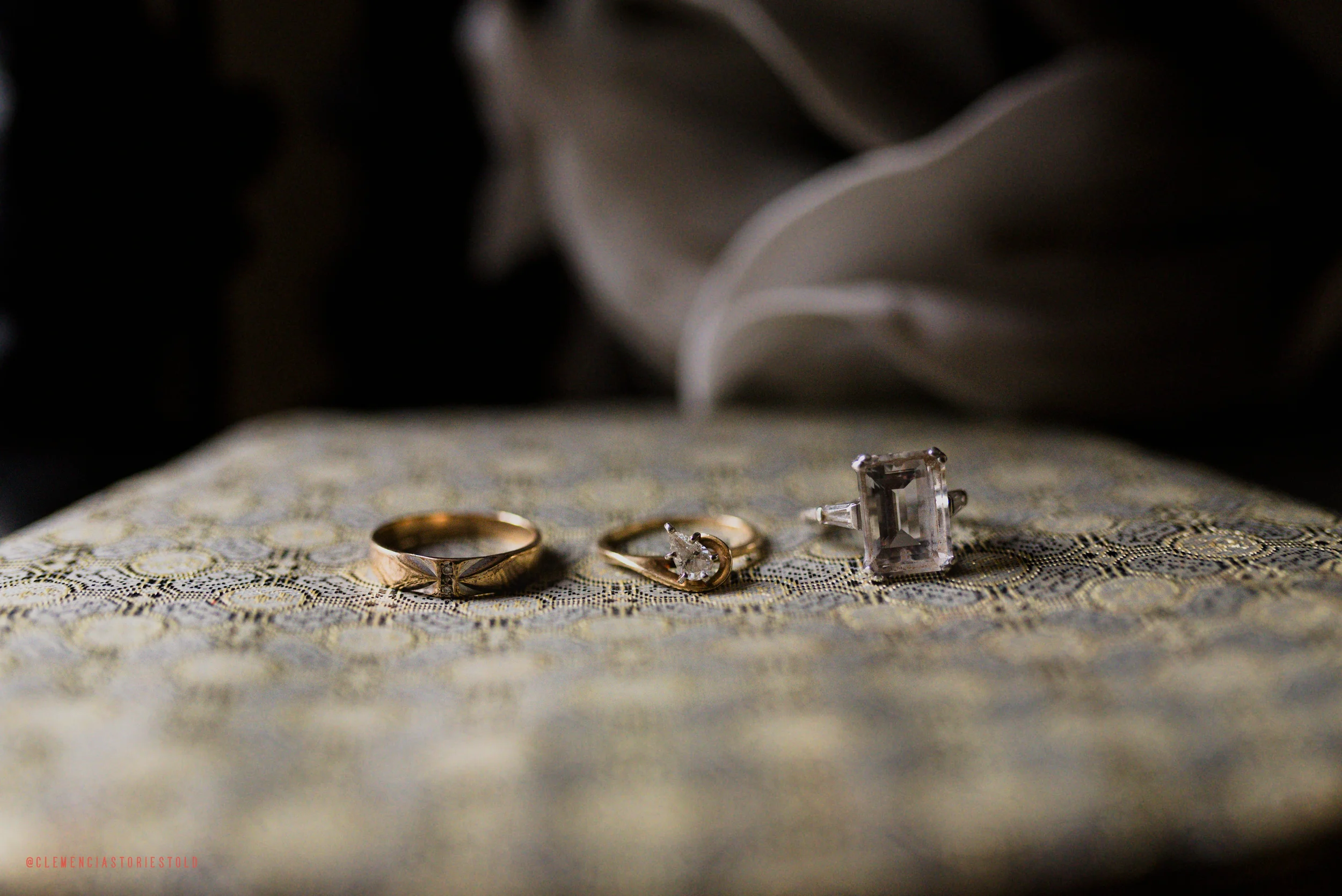 Close-up of three rings and a jewelry box on an ornate fabric surface with white flowers in the background.