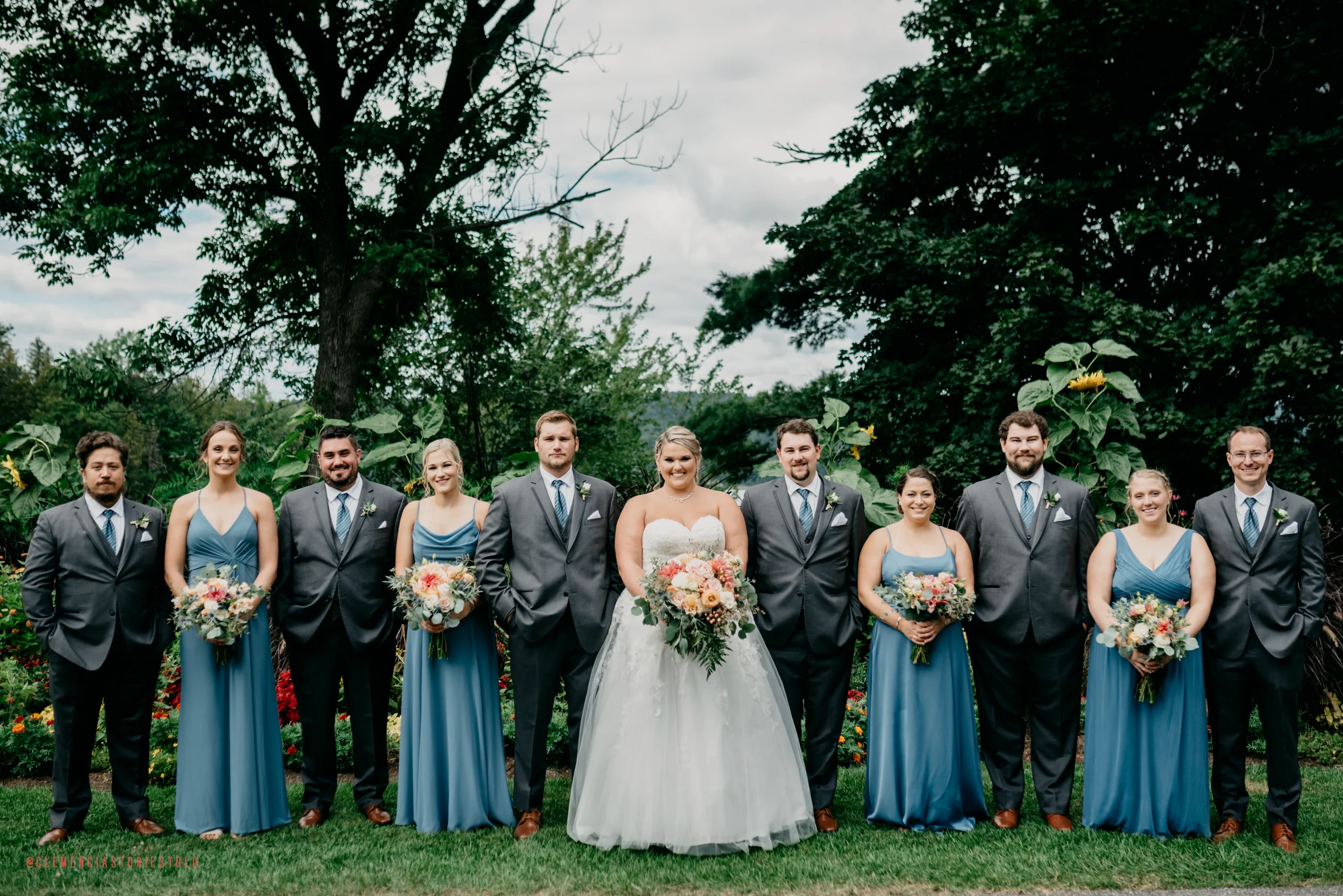 Group of wedding party members standing outdoors on grass with trees in the background, including bride in white gown and bridesmaids in blue dresses, holding bouquets.