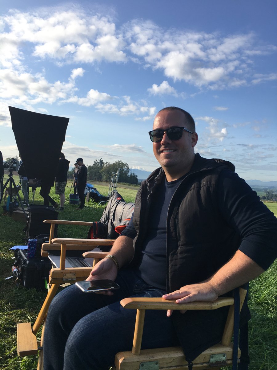 Man sitting on a wooden director's chair outdoors on a film set, holding a smartphone, with film crew and equipment in the background under a partly cloudy sky.