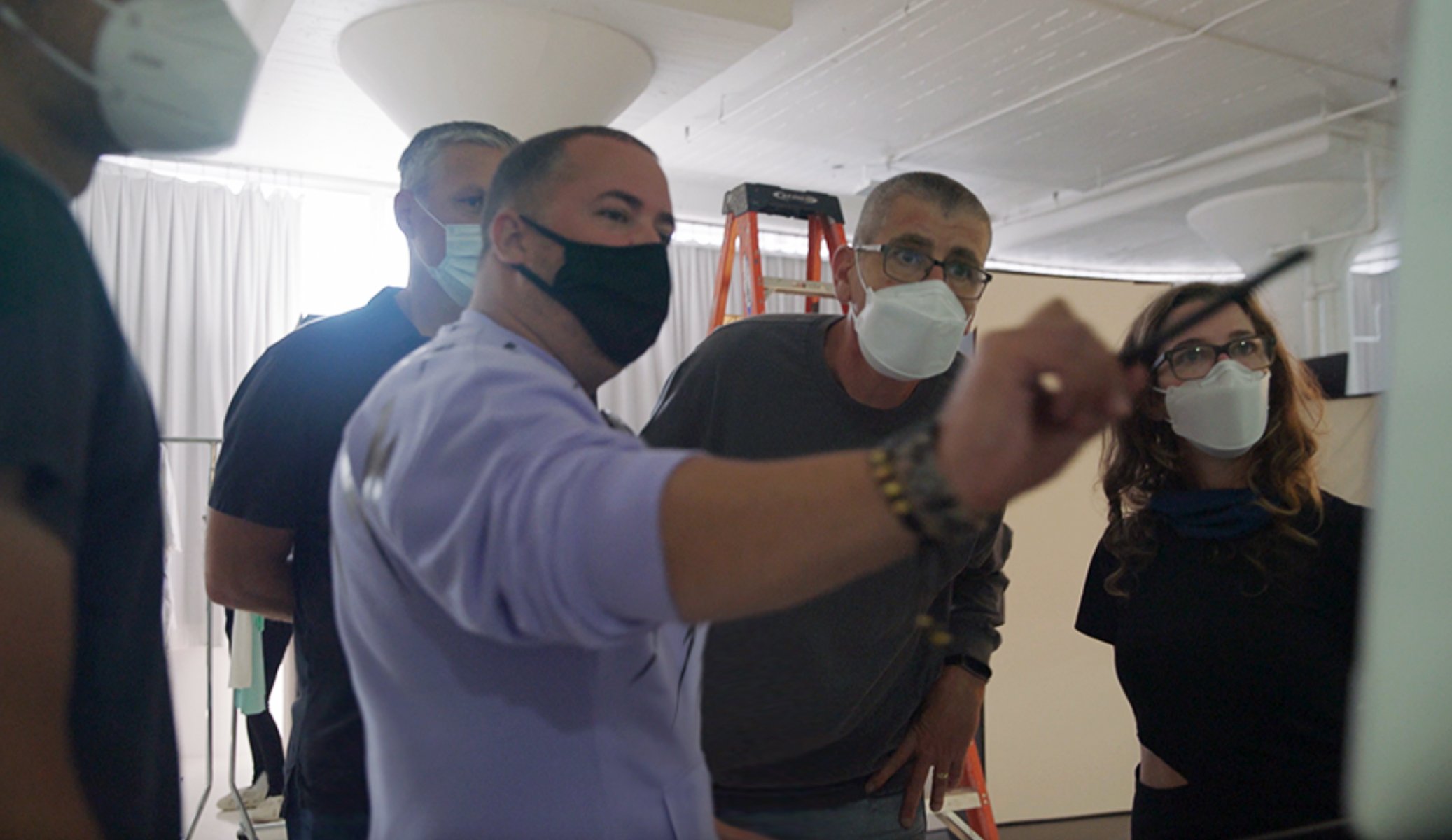 Four people wearing face masks look at a screen or a display, with one person pointing at something on it, in a room with white curtains and a ladder in the background.