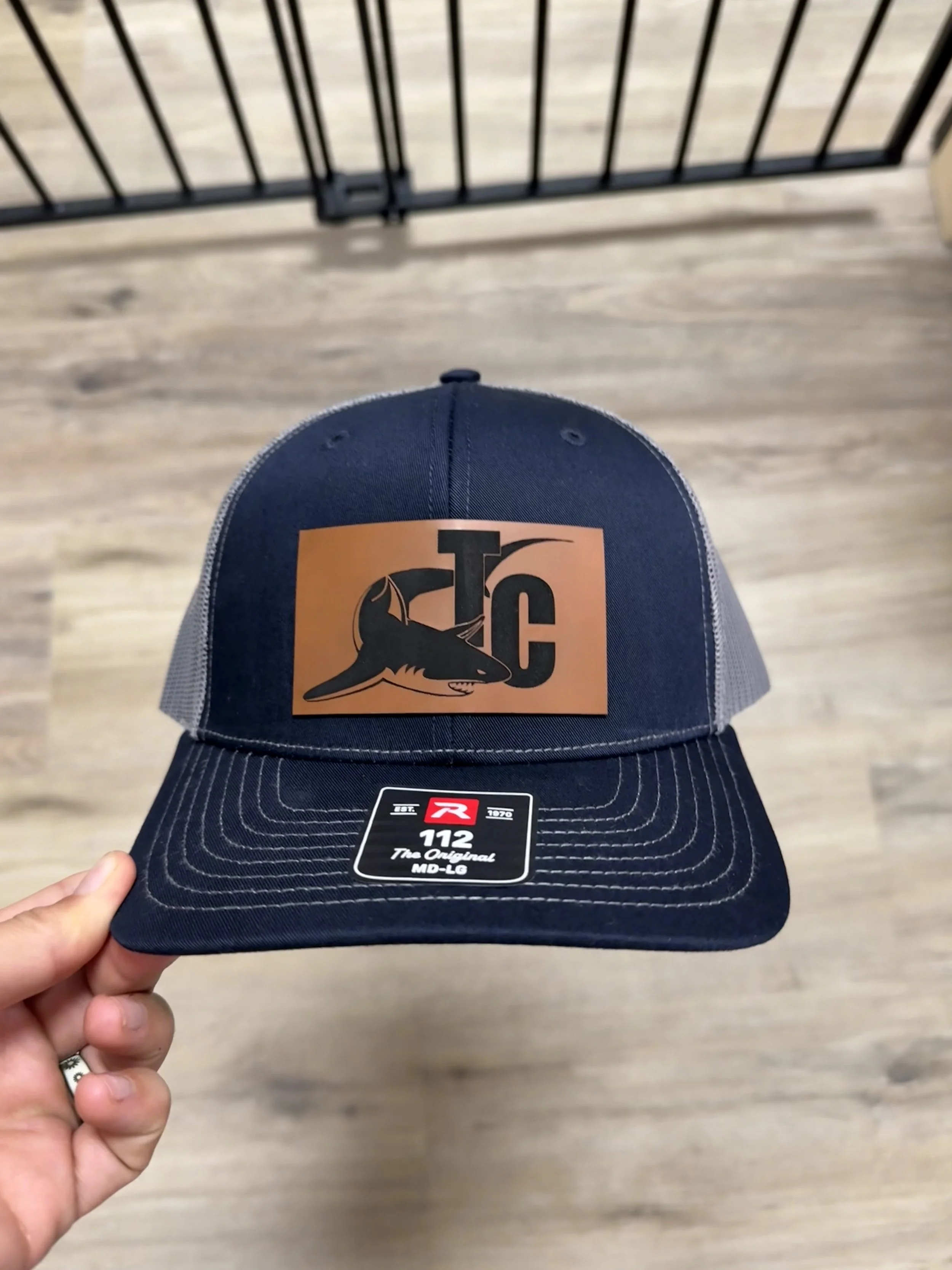 A pink baseball cap with a mountain and tree patch, and the words "Leave Me Alone" embroidered on it, resting on a white surface in front of a blurred window view.