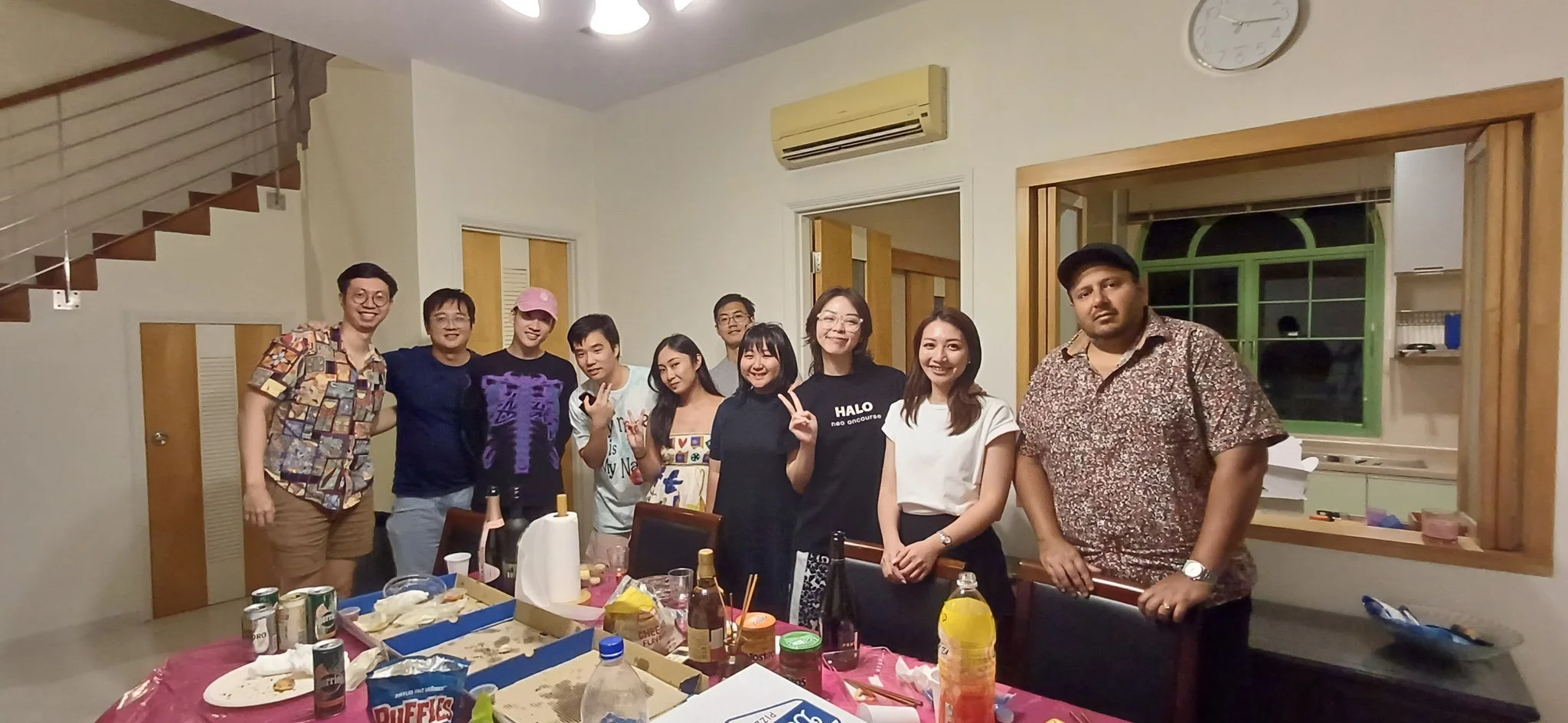 Group of eleven people posing together inside a house, standing behind a table with food and drinks, near a staircase and kitchen window.