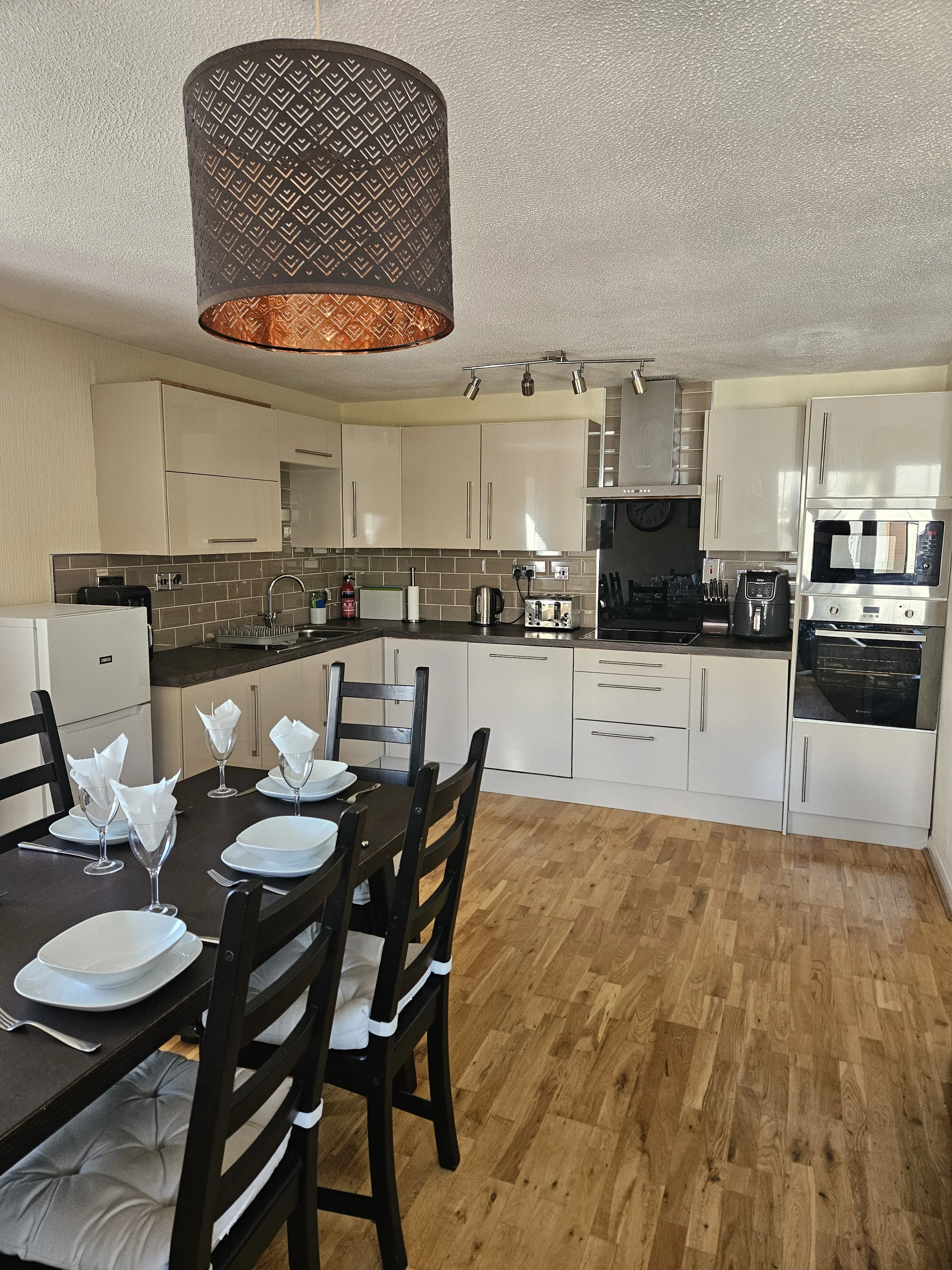 A modern kitchen with white cabinets, a dark countertop, a microwave, an oven, and a dining table set with white plates and napkins, featuring wooden flooring and a ceiling light fixture.