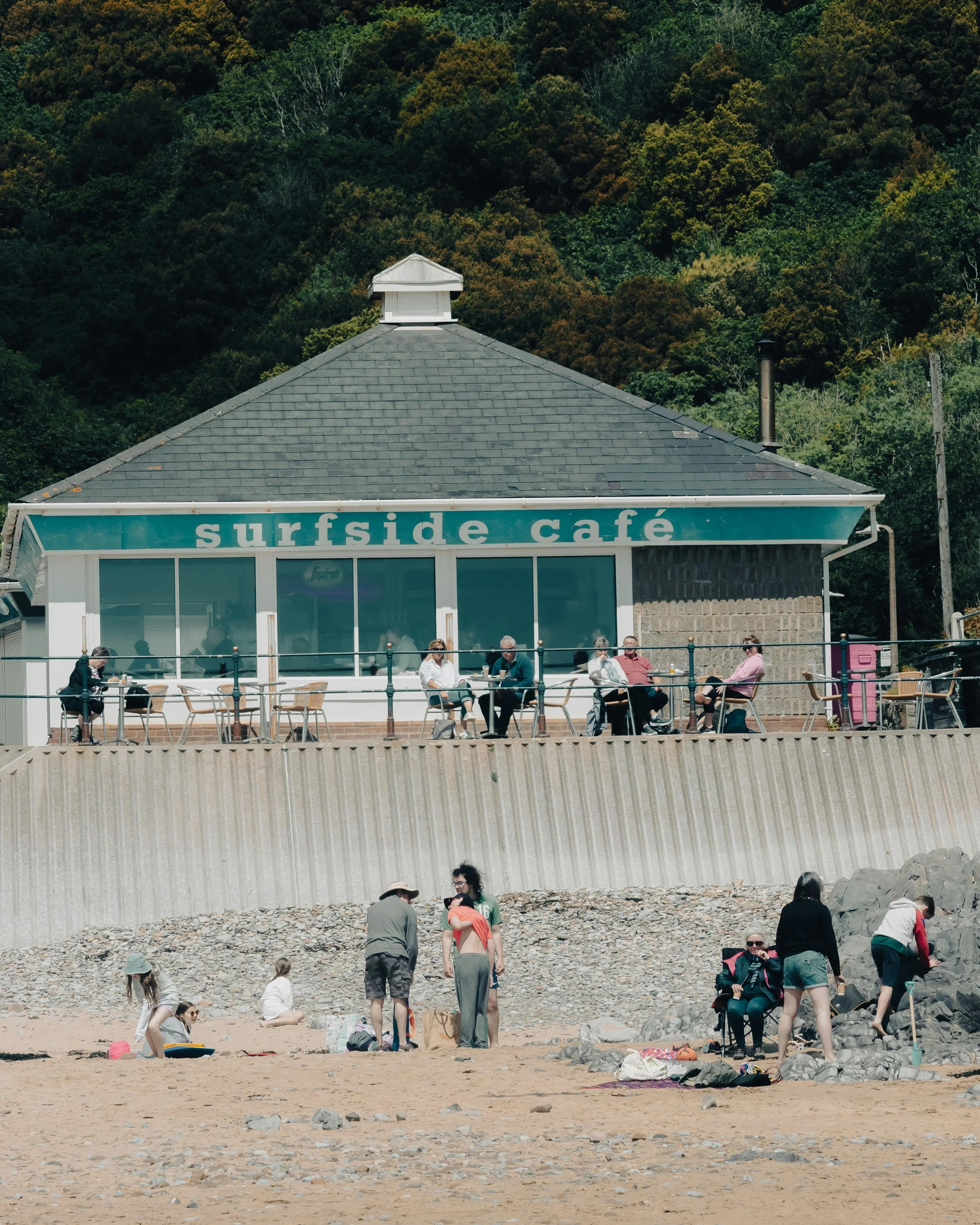 People sitting at tables outside a café with a forested hillside in the background, and others walking or sitting on a rocky beach in front.