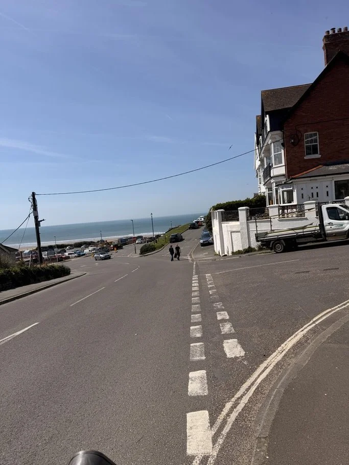 A sloped street leading to the beach, with cars parked along the sides and two people walking toward the ocean in the distance. A house is on the right side of the street, and the sky is clear and blue.