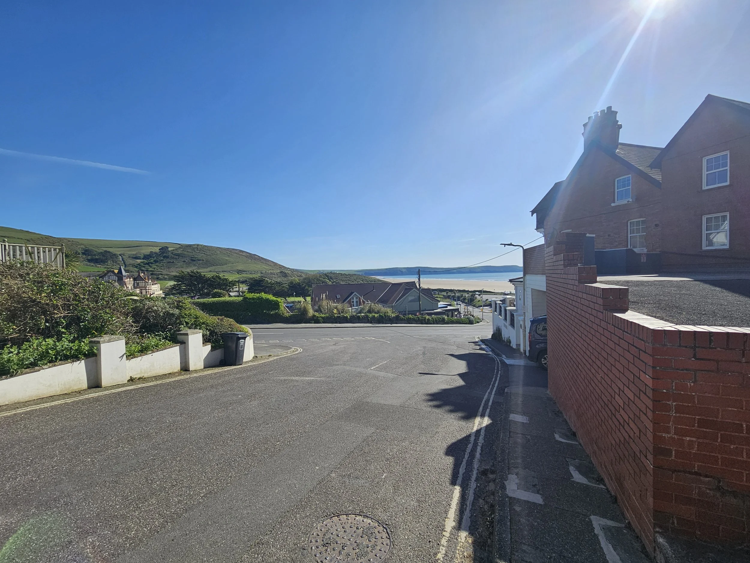 View of a coastal town with houses, hills, and a beach, under a bright blue sky with the sun shining and a contrail visible.