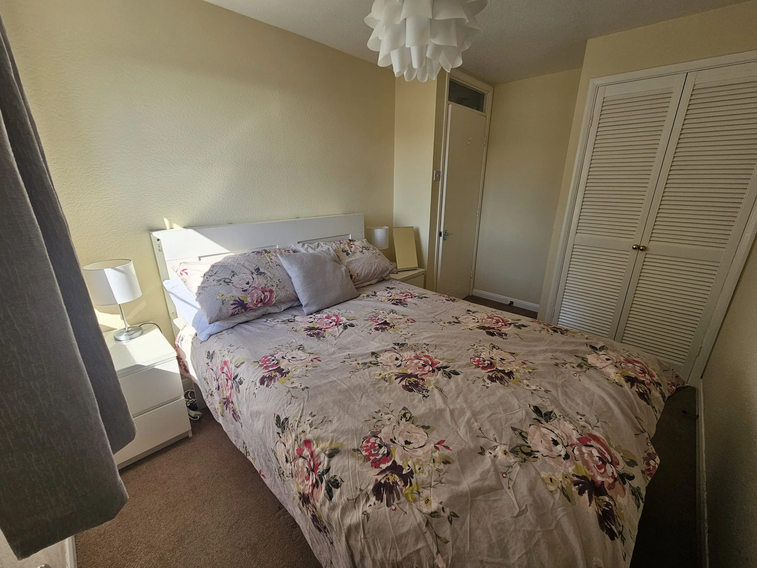 A bedroom with a bed covered in floral bedspread, two pillows, bedside table with a lamp, a closet with louvered doors, and a door next to a small window. Light from the window casts a shadow on the bed.