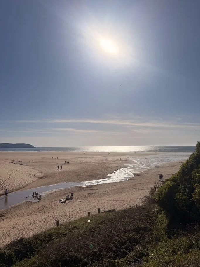 A sunny beach scene with people walking and relaxing on the sandy shore, small streams flowing into the ocean, and a calm sea under a bright blue sky with the sun shining.