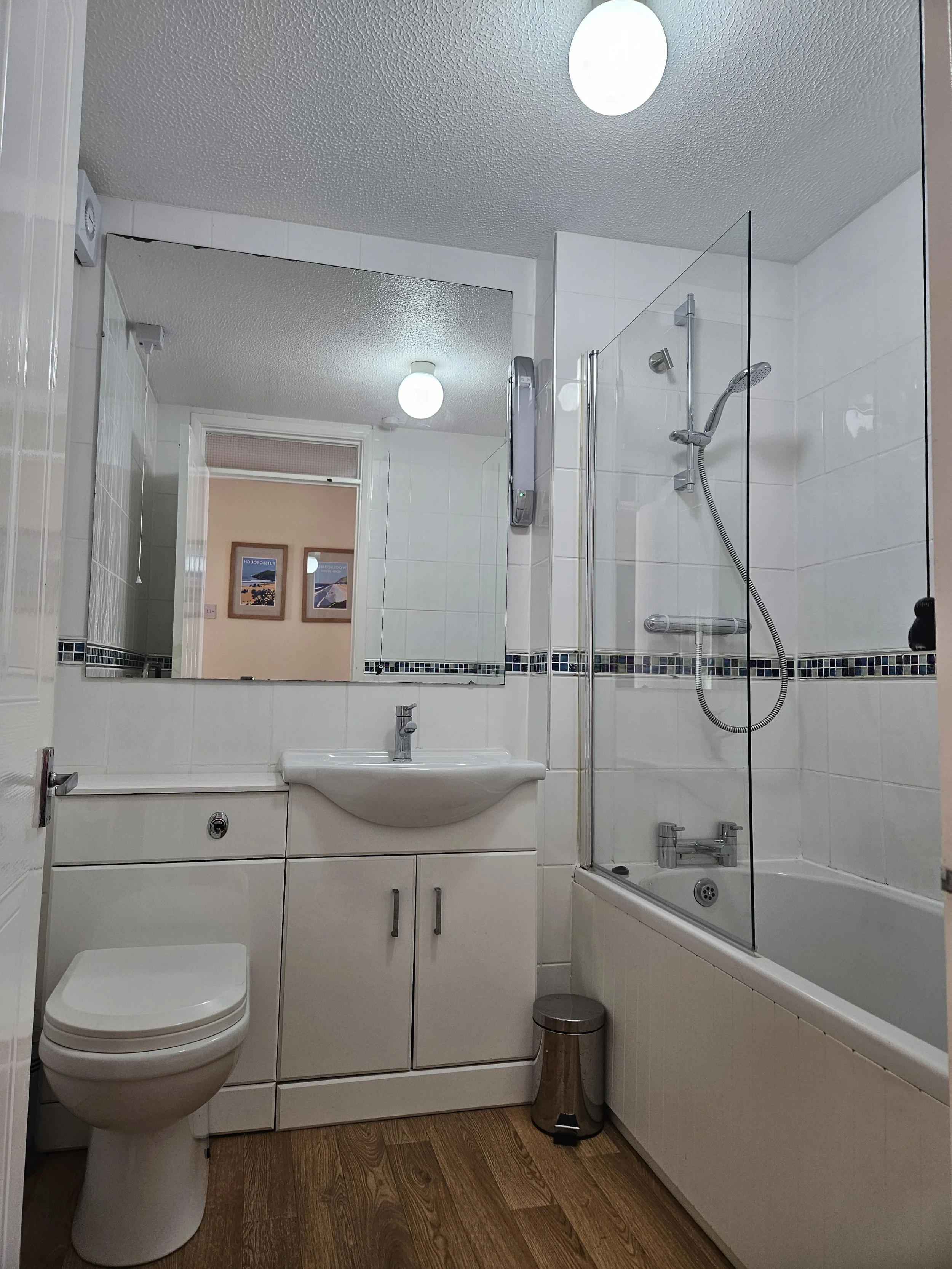 A bathroom with white tiled walls, a toilet, a sink with a mirror above, a bathtub with a glass shower screen, and a wooden floor.