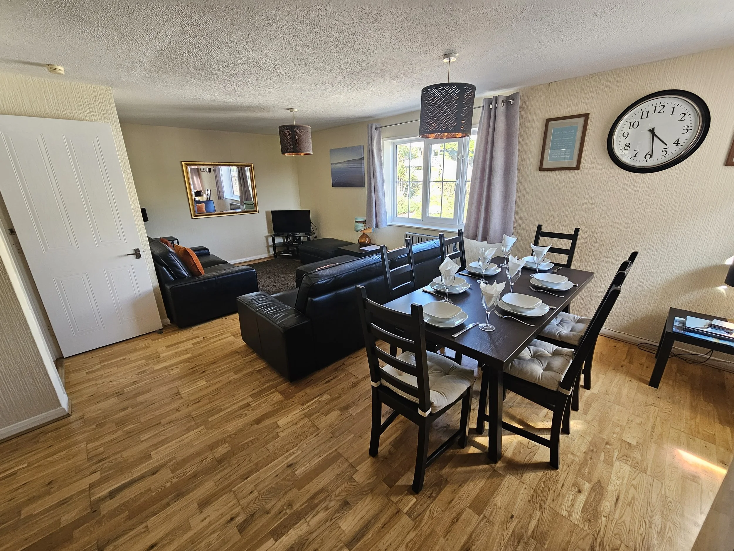 A living room and dining area with a black dining table set for dinner, a black leather sofa, a small television with a stand, a window with curtains, framed wall art, and a wall clock showing 4:31.