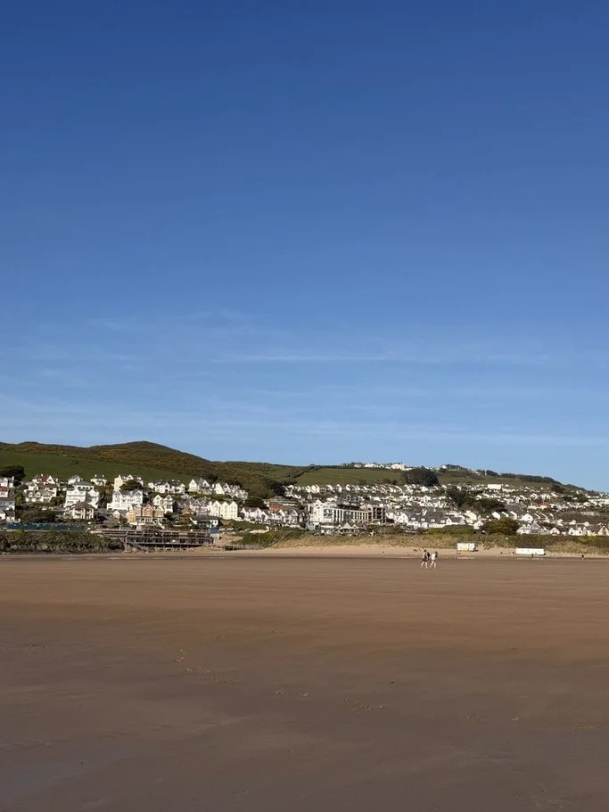 Beach with a wide sandy shoreline under a clear blue sky, with a small group of people walking in the distance and a hillside with white houses in the background.