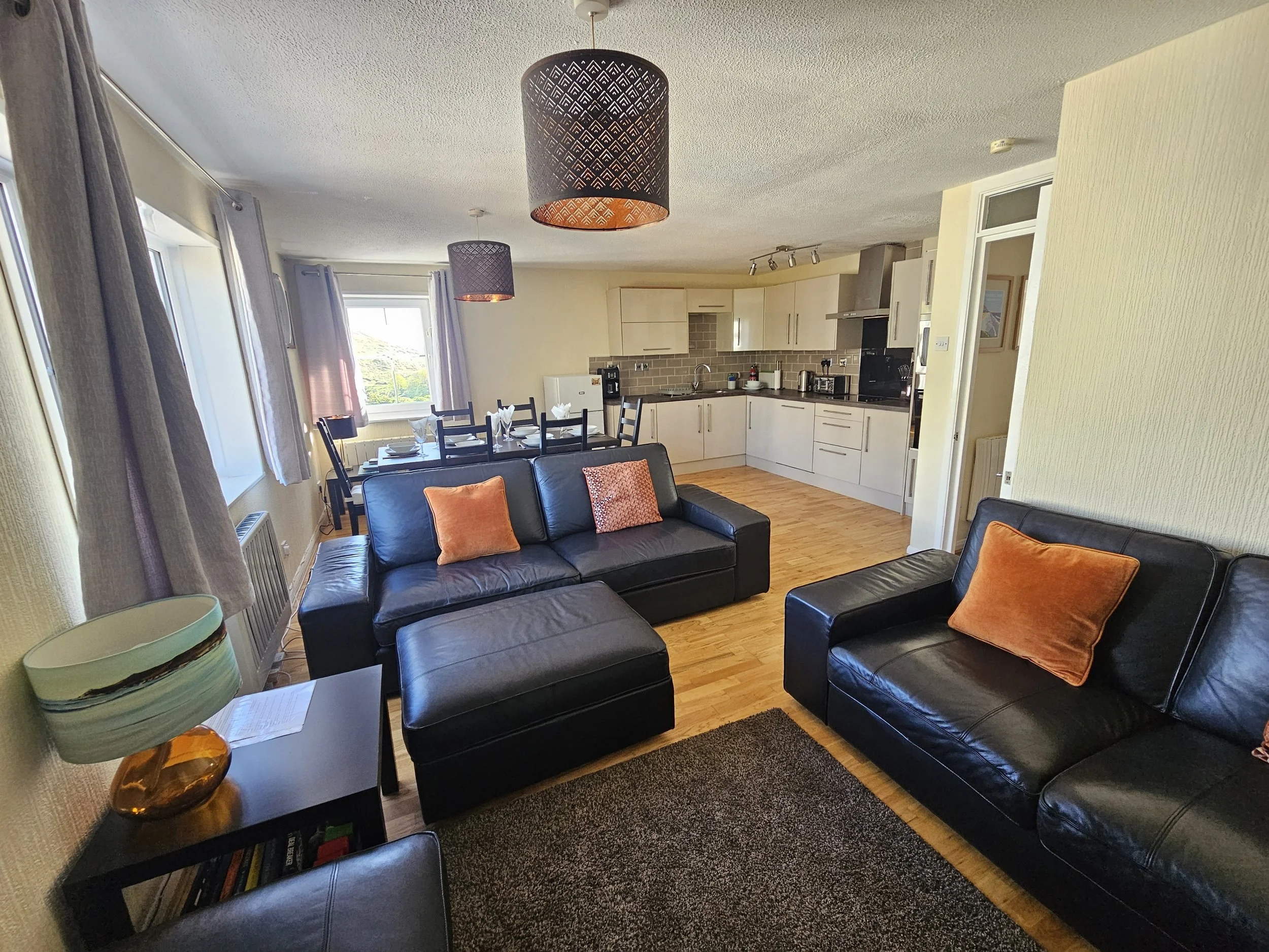 A living room with black leather sofas, orange pillows, a black table, a lamp, and a view of the kitchen in the background, with beige walls and wooden flooring.