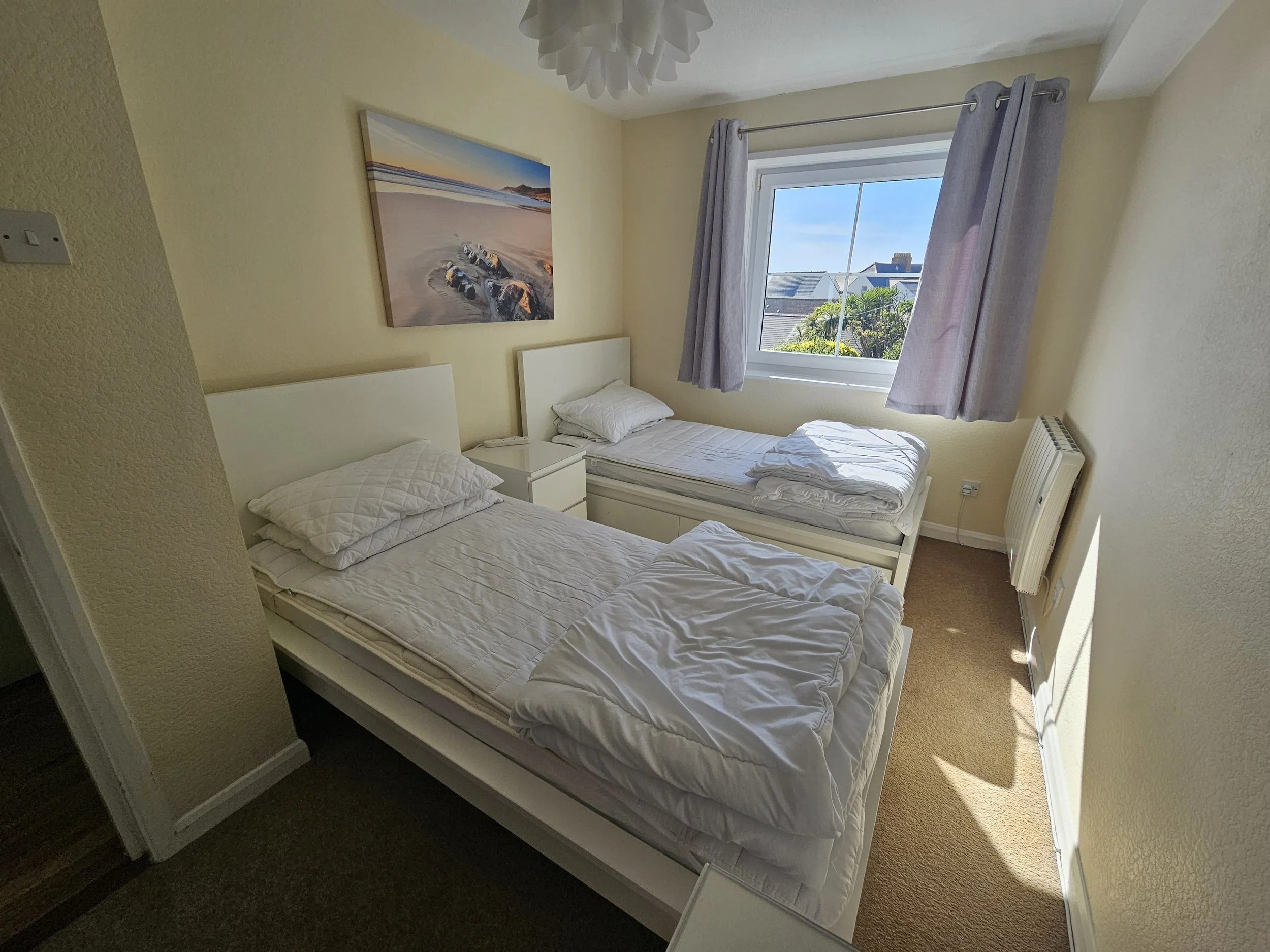 A bedroom with two single beds, a window with purple curtains, and a painting of a beach scene on the wall.