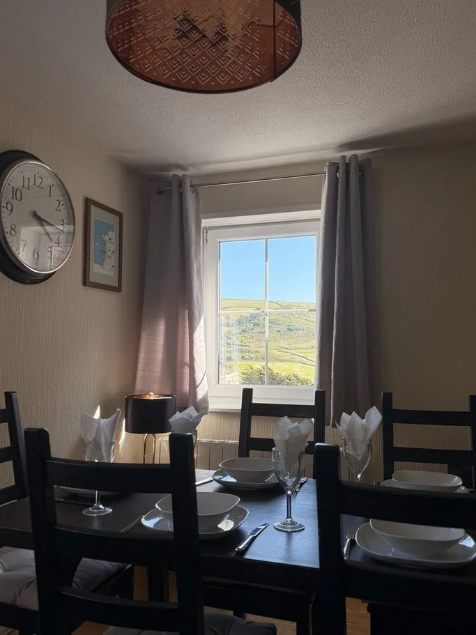 A dining room with a dark wooden table set with plates, wine glasses, and folded napkins. Four black chairs are around the table. There is a window with light curtains, revealing a view of green hills and blue sky outside. A wall clock and a framed p
