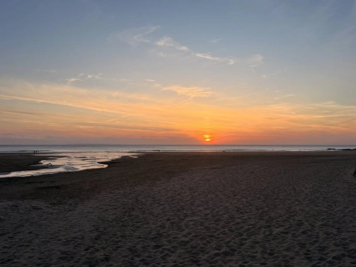 Sunset over a quiet beach with a calm ocean, some small streams in the sand, and a few distant people along the shoreline.