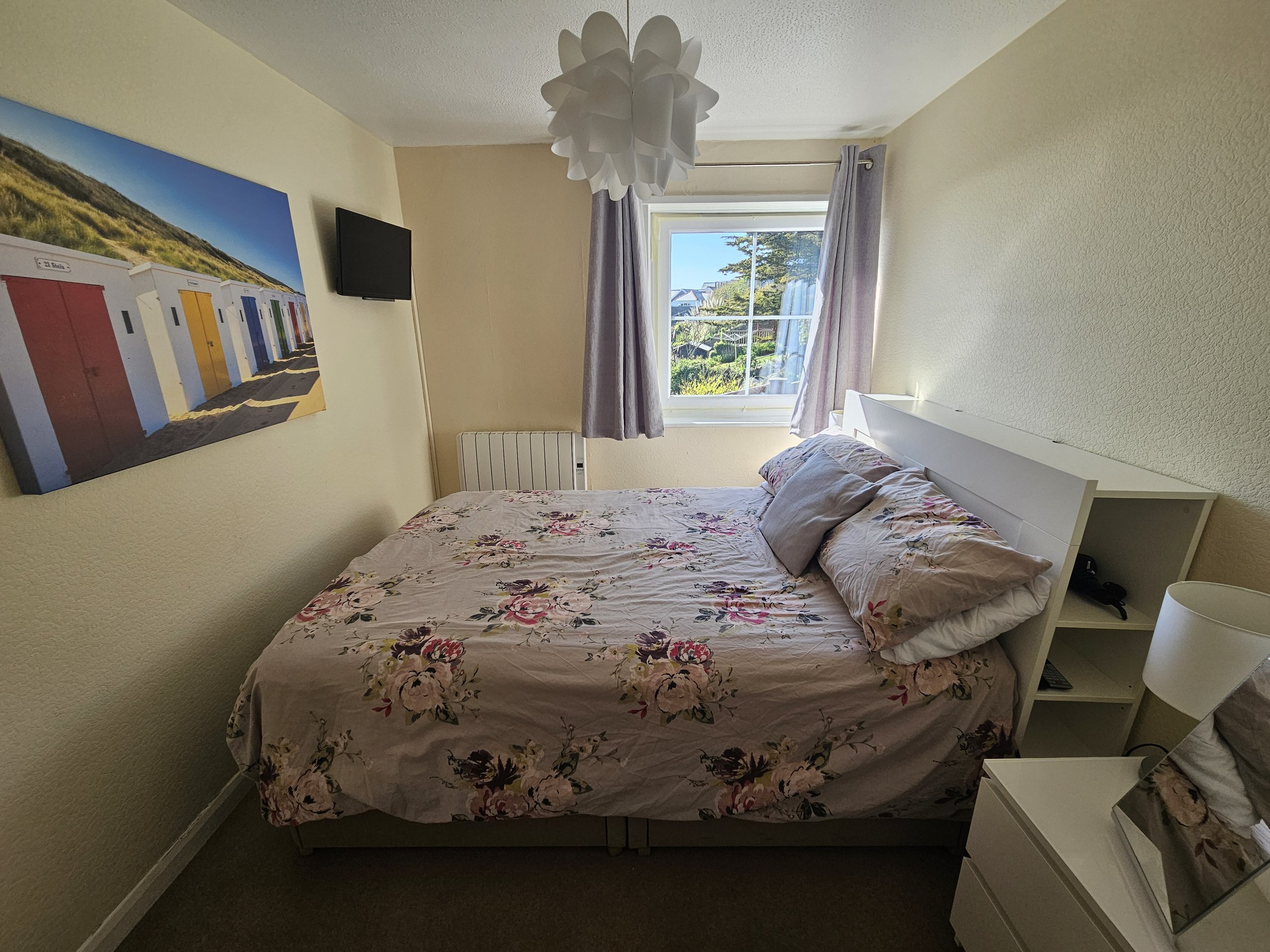 A bedroom with a bed covered in floral sheets, a white bedside table with a lamp, a wall-mounted TV, a window with light purple curtains, a painting of colorful beach huts, and a white ceiling light fixture.