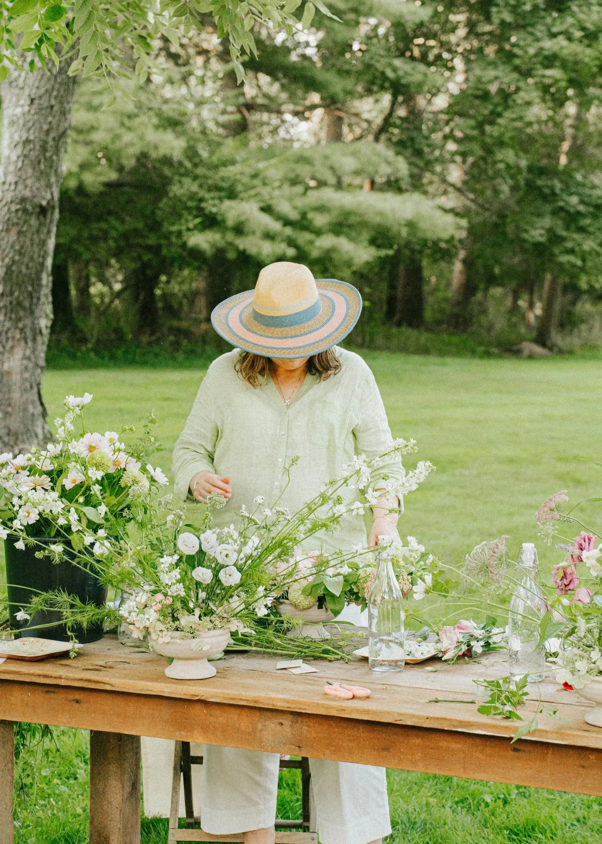 A woman wearing a wide-brimmed straw hat and a light-colored shirt arranging flowers on a wooden table outdoors in a grassy area with trees in the background.