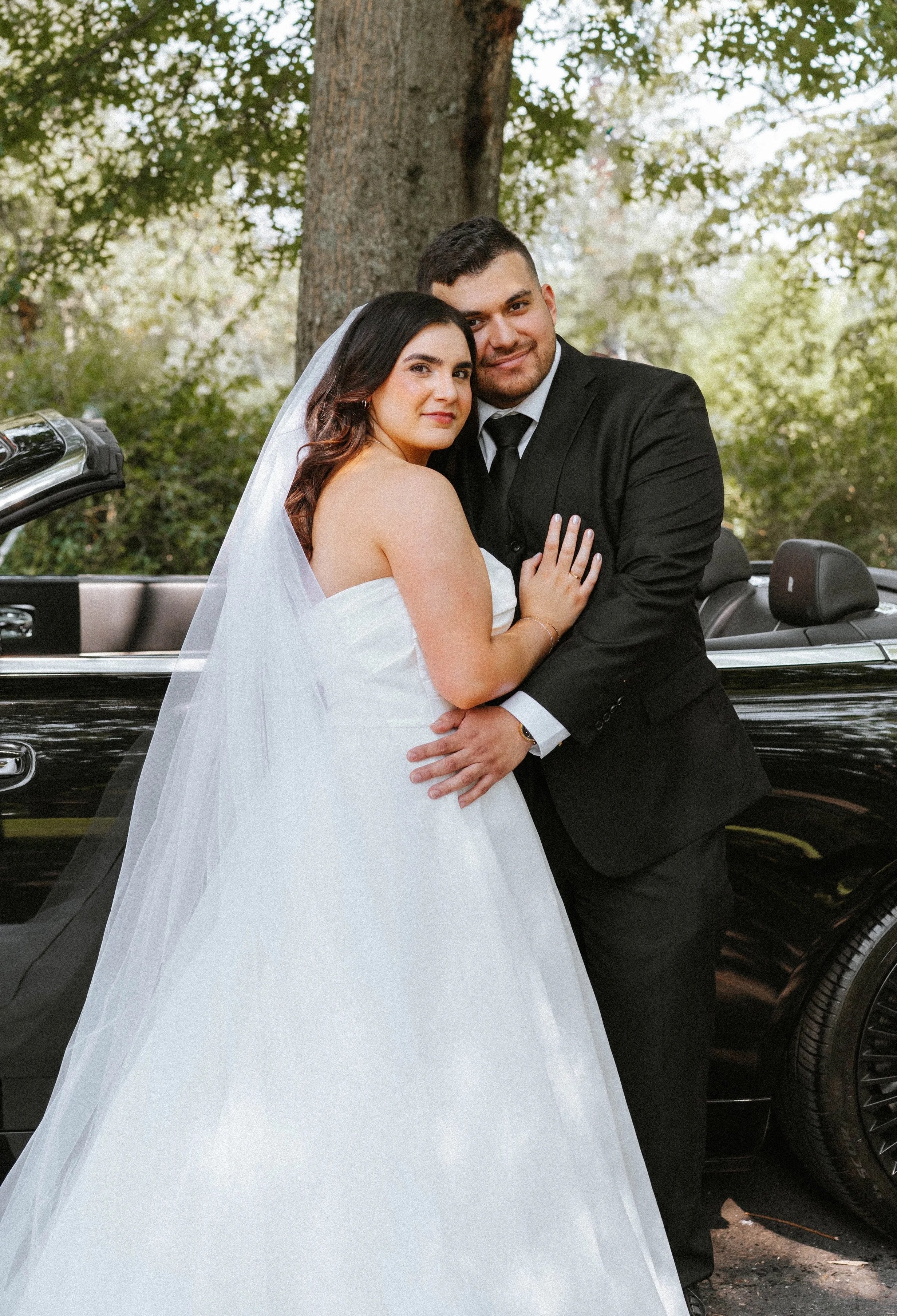 A bride and groom in wedding attire standing close together outdoors in front of a black convertible car. The bride is wearing a white wedding gown with a veil, and the groom is dressed in a black suit with a white shirt and black tie. They are embra