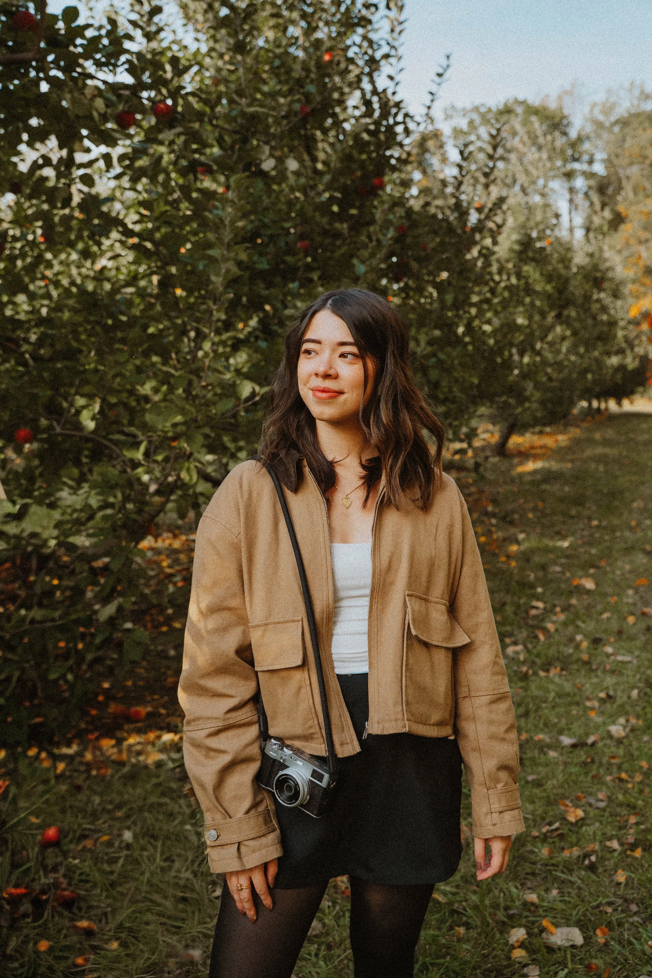 A young woman with shoulder-length wavy hair, wearing a tan jacket, white top, black skirt, and tights, standing outdoors in an apple orchard during fall, with apple trees in the background, some apples visible on the trees and on the ground. She has a camera hanging around her neck and is gazing softly to the side.