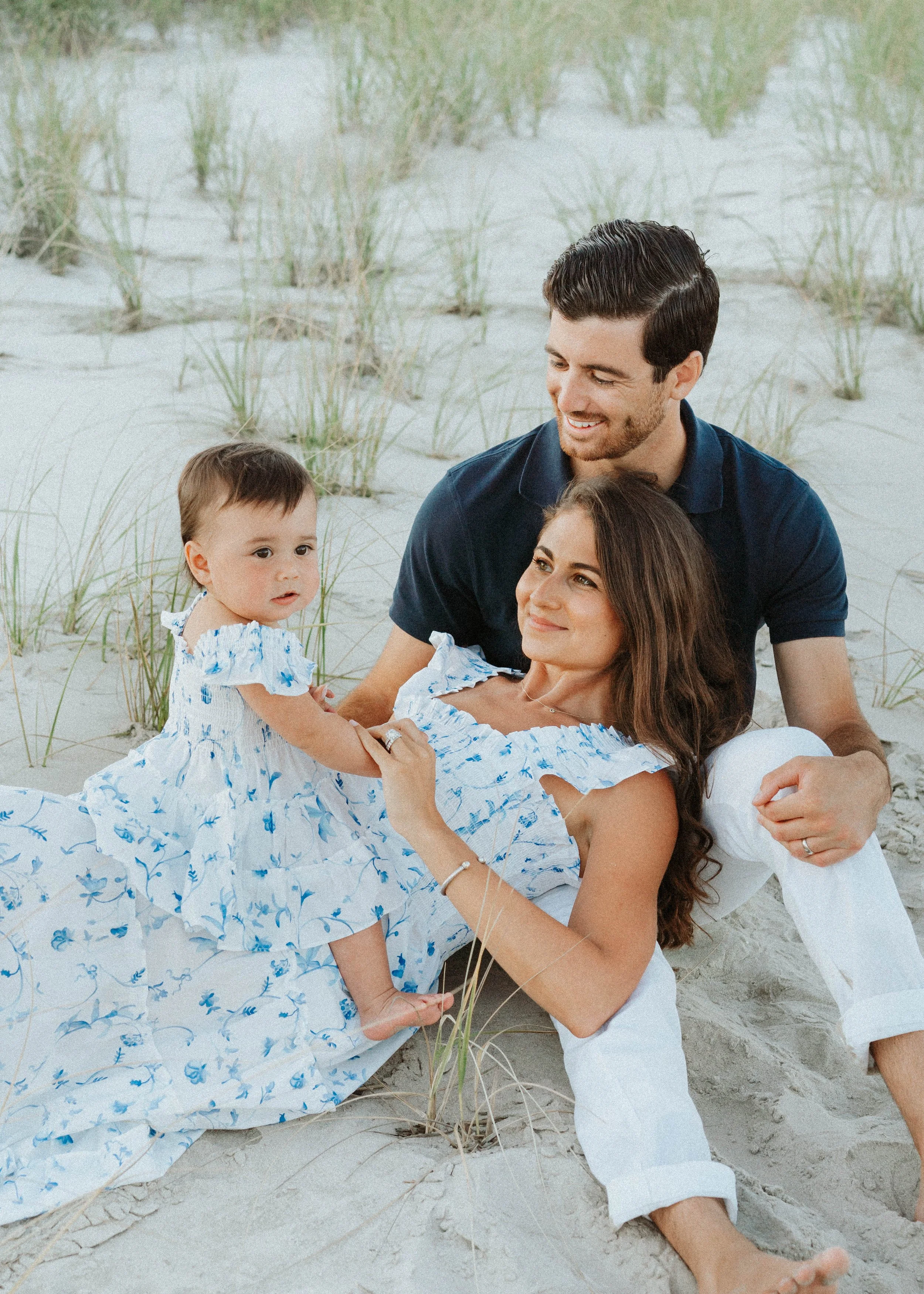 A family of three, including a woman, a man, and a young girl, sitting on the sand at the beach with grass in the background. The woman is lying down, holding the girl, while the man is sitting behind her, smiling and looking at the girl.