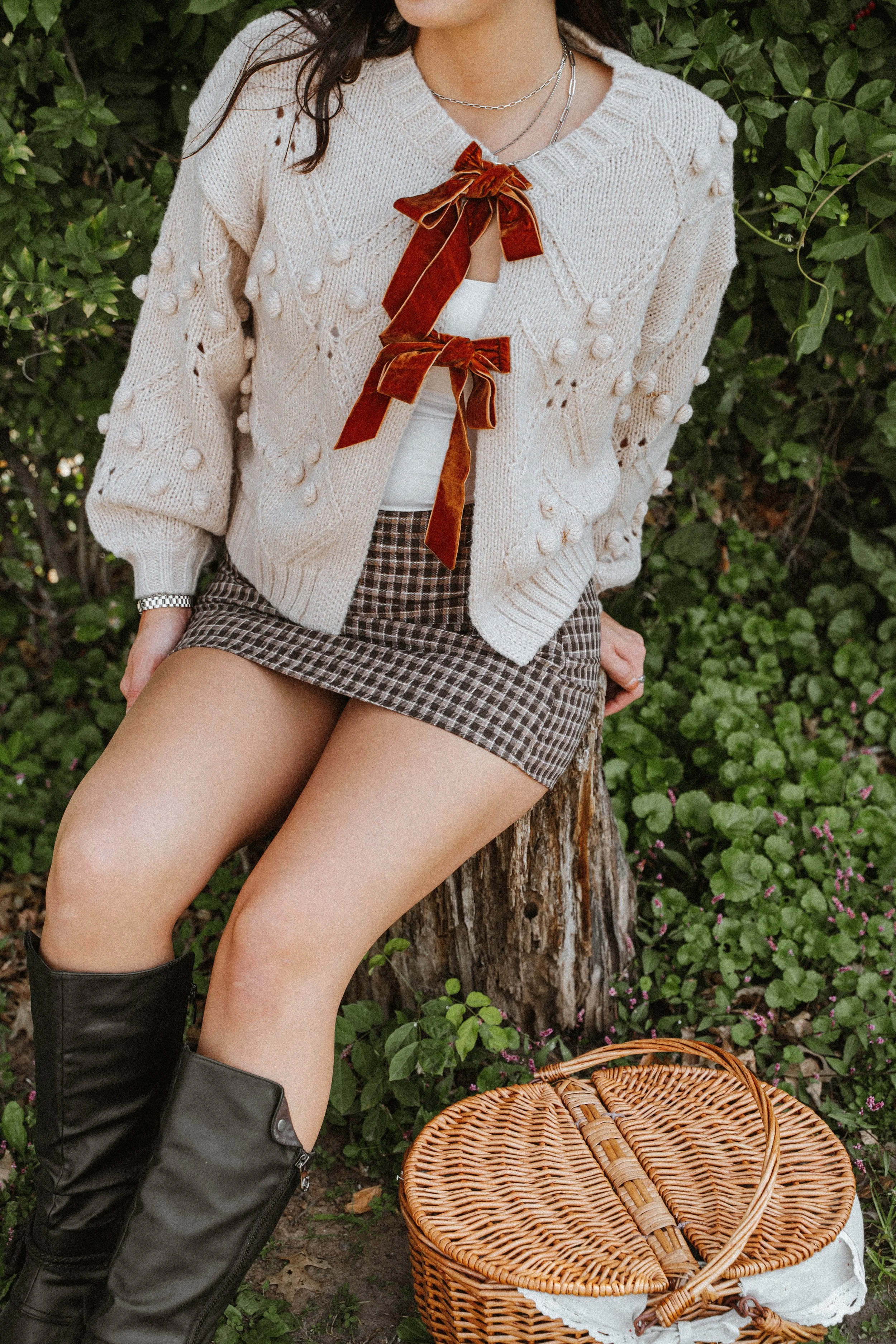 A woman sitting outdoors on a tree stump, wearing a beige cardigan with pom-pom details, a white top, a plaid mini skirt, and black boots. There is a wicker basket on the ground nearby.