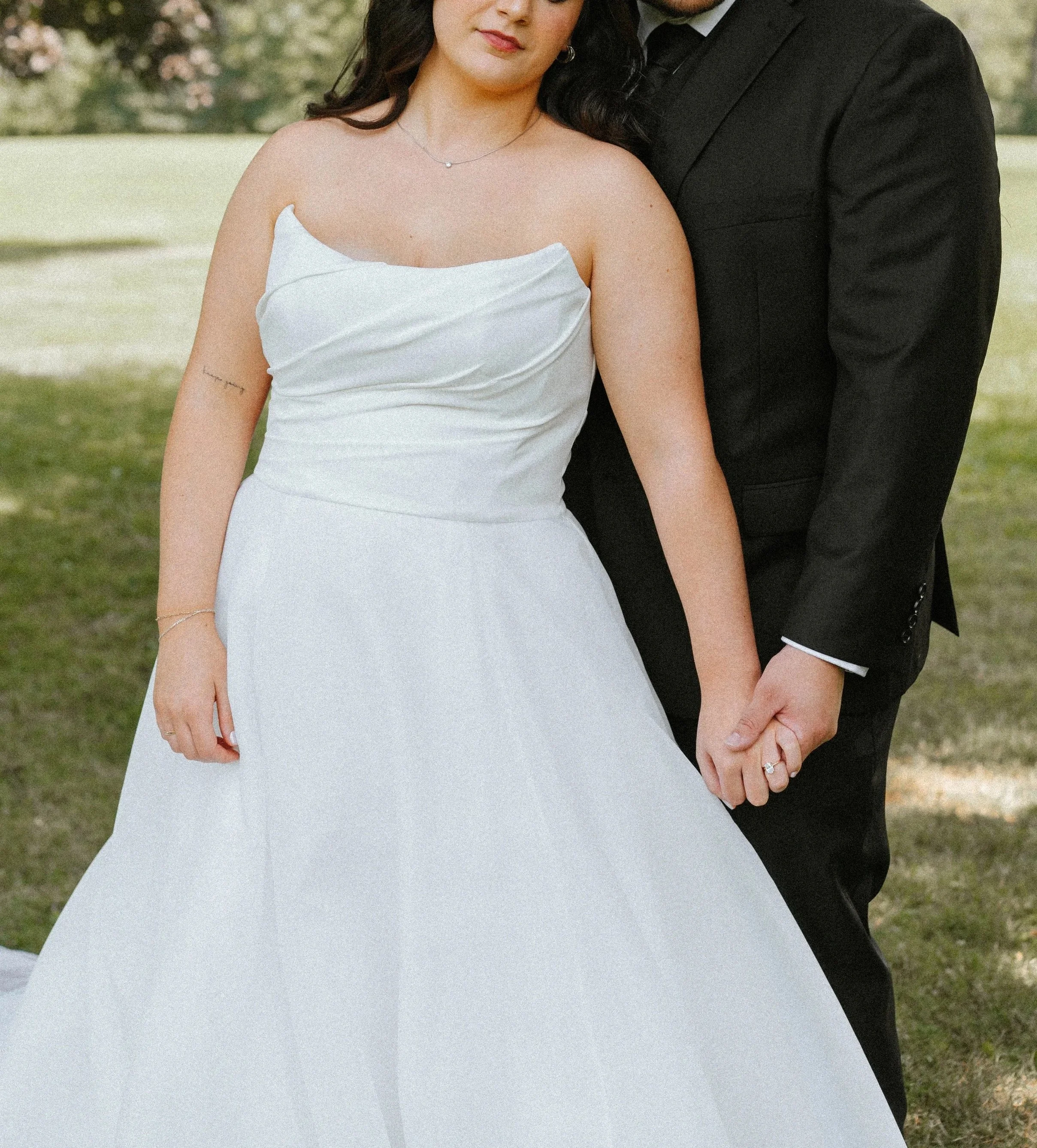 A bride and groom holding hands outdoors, with the bride wearing a white wedding dress and the groom in a black suit, standing on grass with trees in the background.
