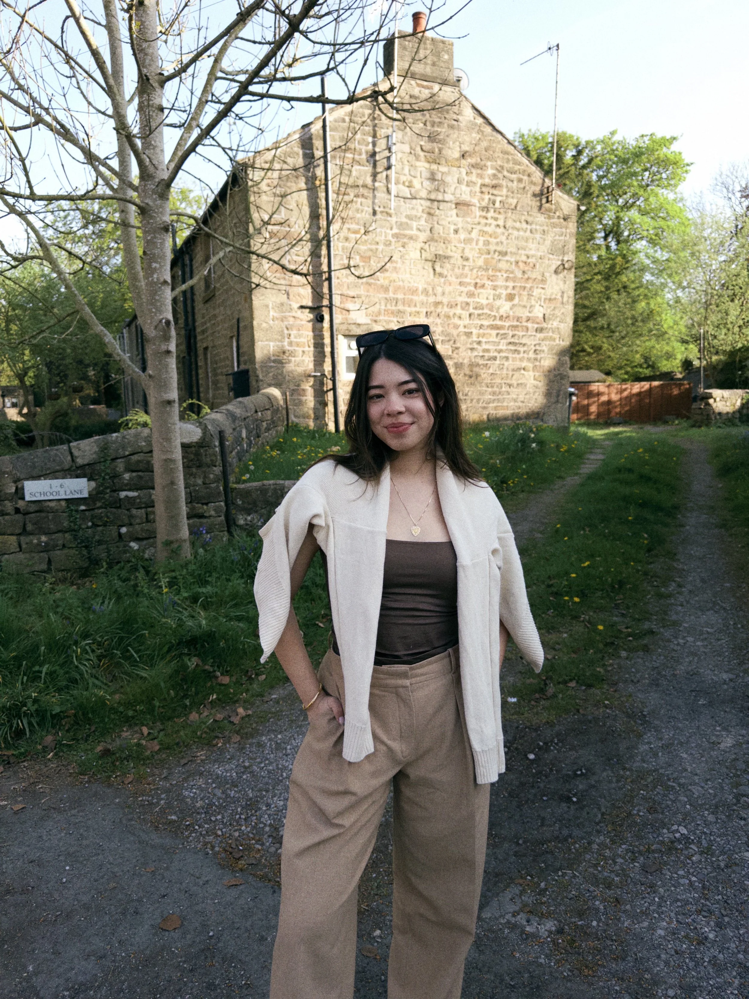A woman standing outdoors on a dirt path in front of a stone house with trees and greenery around, wearing a brown top, beige pants, a cream sweater draped over her shoulders, and sunglasses on her head.