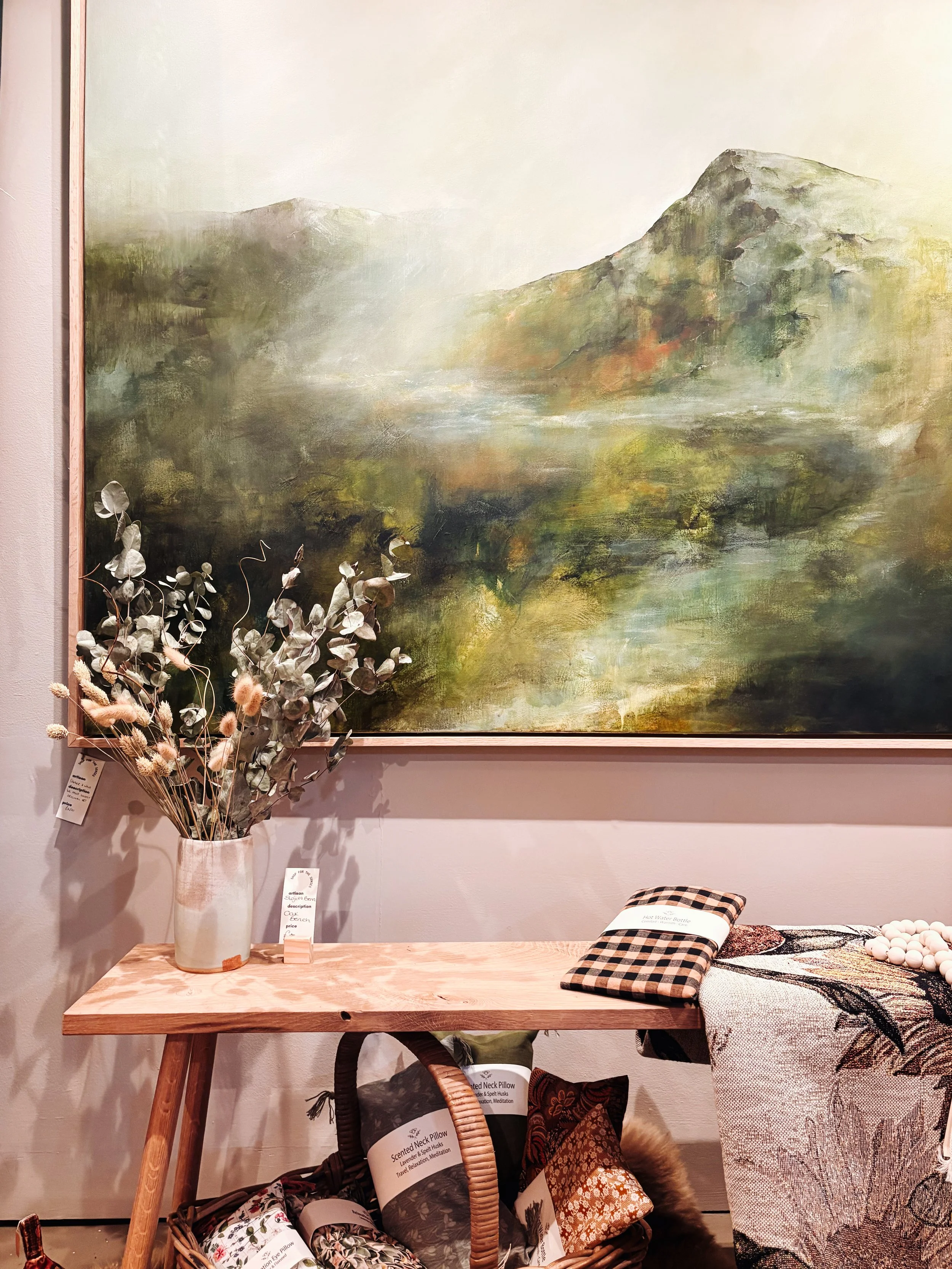 A corner of a room with a large landscape painting on the wall, a wooden table with a vase of dried flowers, a checkered pillow, and various textiles and pillows underneath.