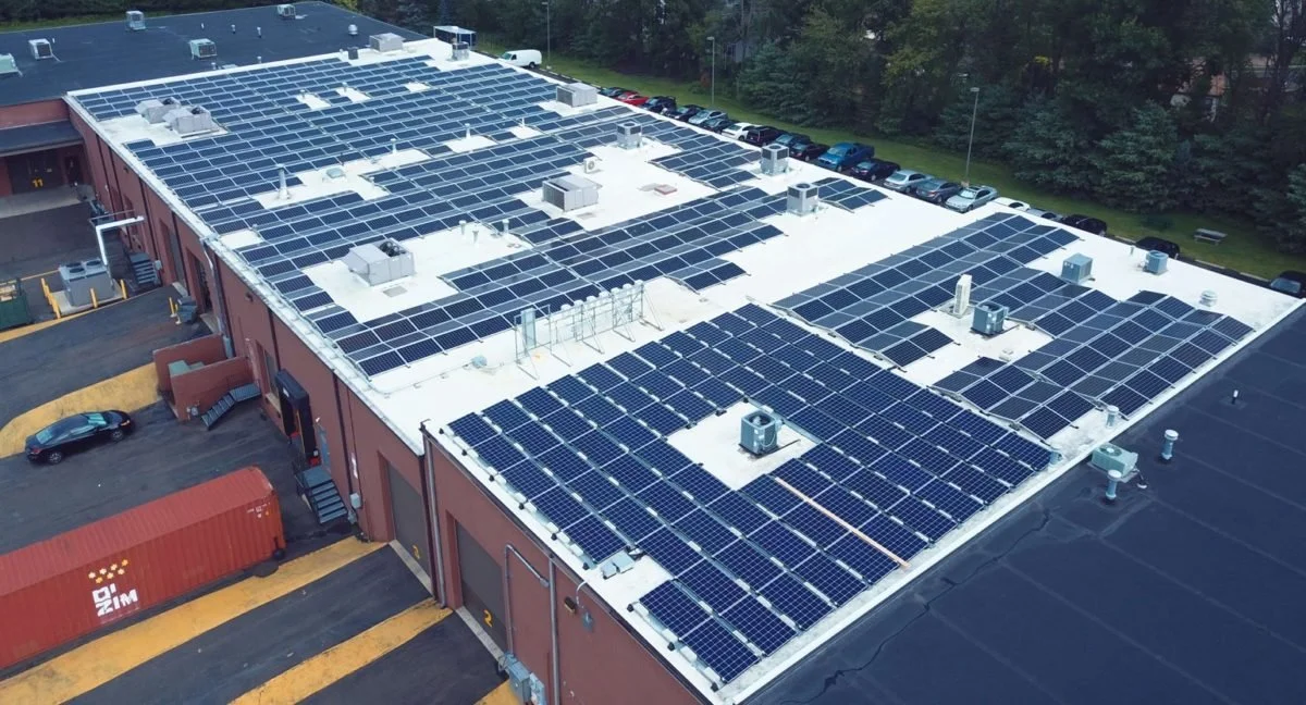 Aerial view of a building rooftop covered with numerous solar panels, with parked cars and trees in the background.